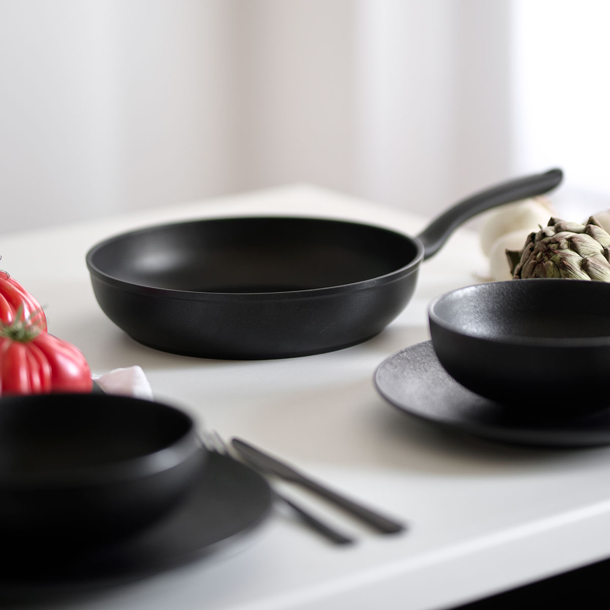 Czarna patelnia, dwie miski i talerze na białym stole. Obecne są również warzywa.