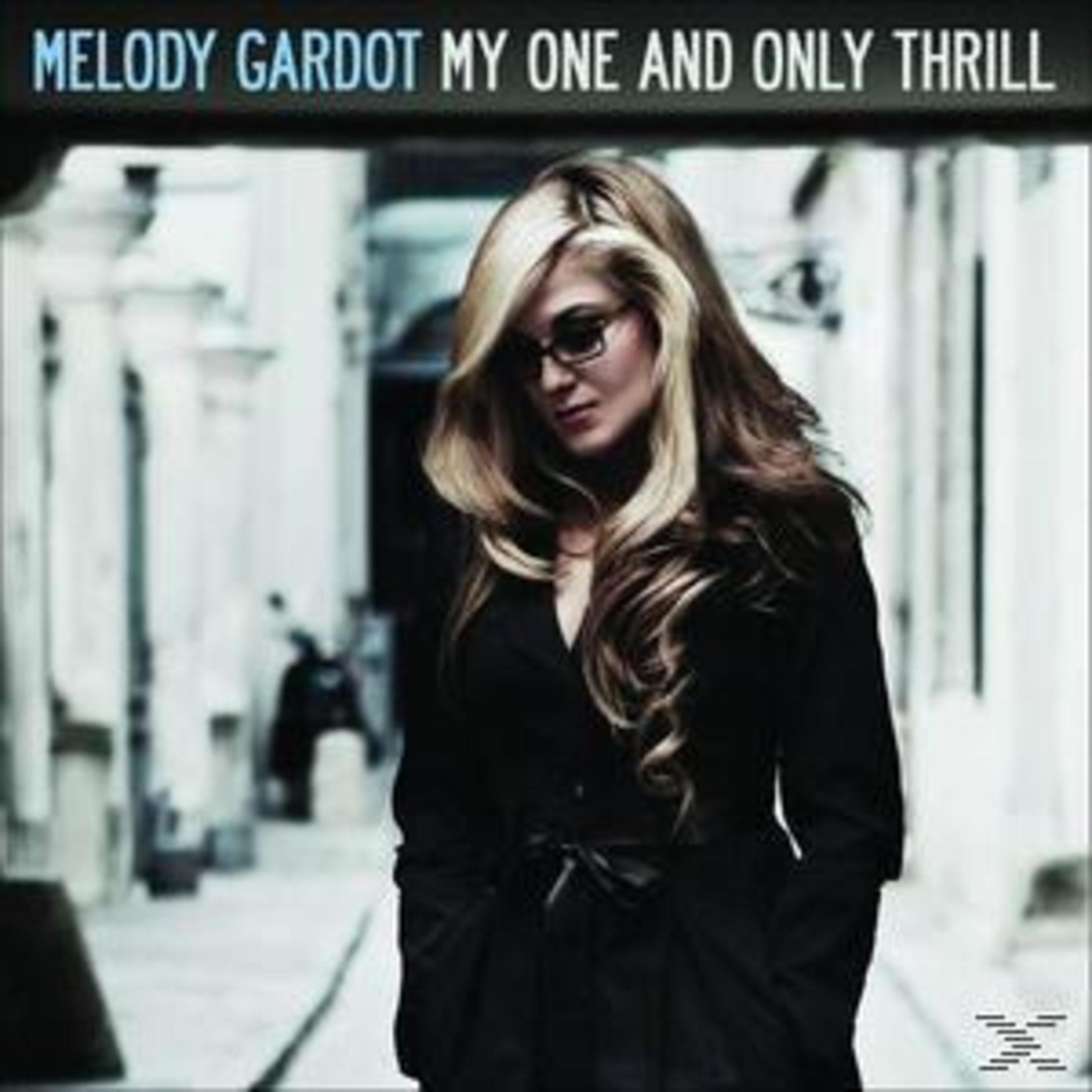 Melody Gardot in posa in un abito nero, con una scena di strada sfocata sullo sfondo.