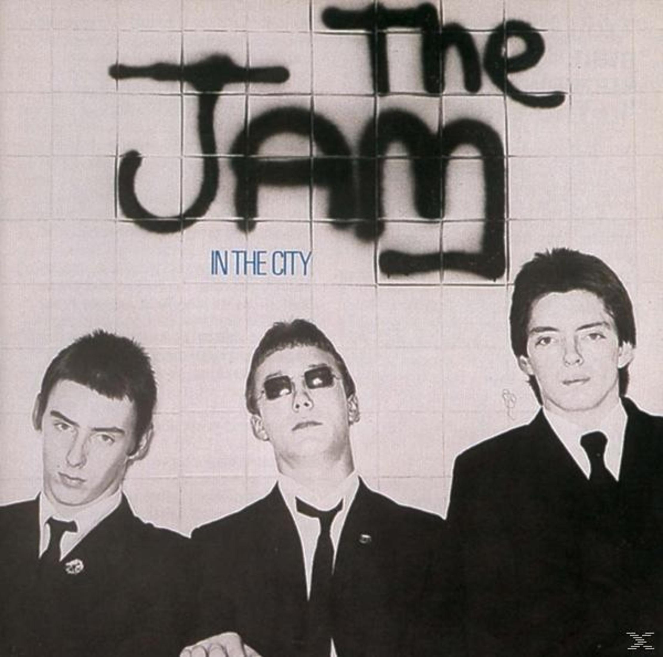 Copertina dell'album: Tre uomini in giacca e cravatta, testo nero: The Jam, In The City, stile graffiti su un muro bianco.