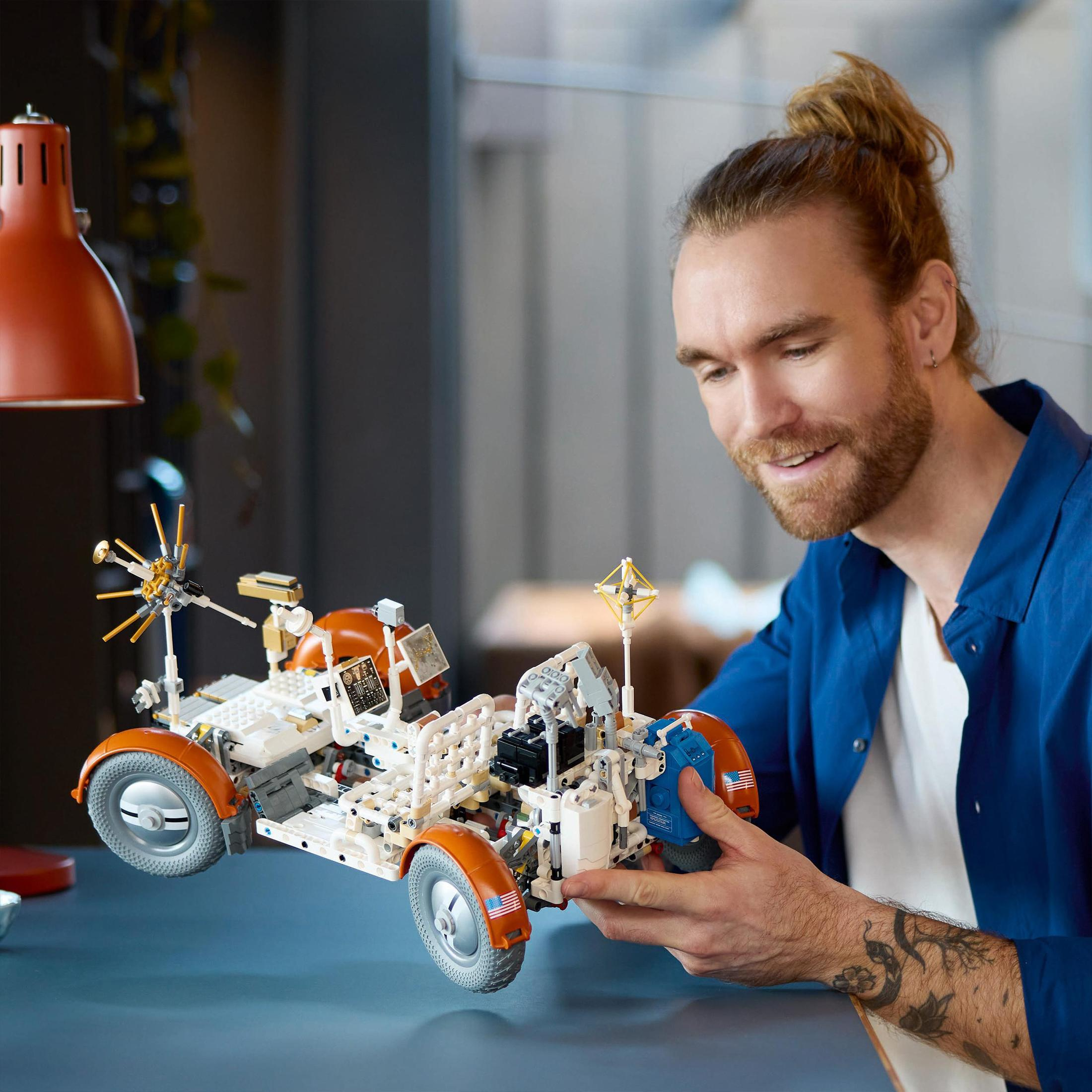 Un uomo sorride a un modello Lego di rover marziano su una scrivania, con illuminazione arancione.