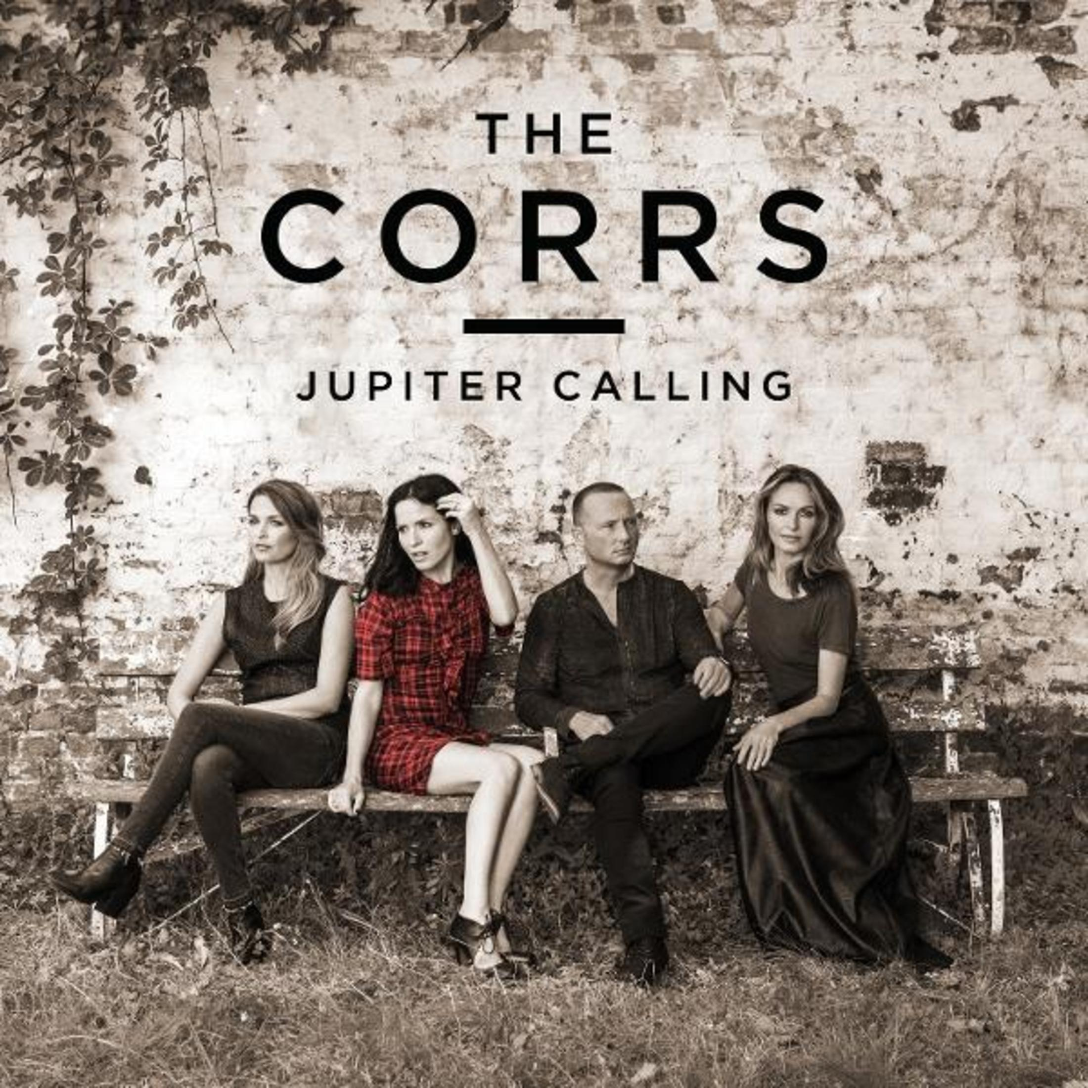 La band The Corrs seduta su una panchina. Lo sfondo è un muro di pietra e erba.