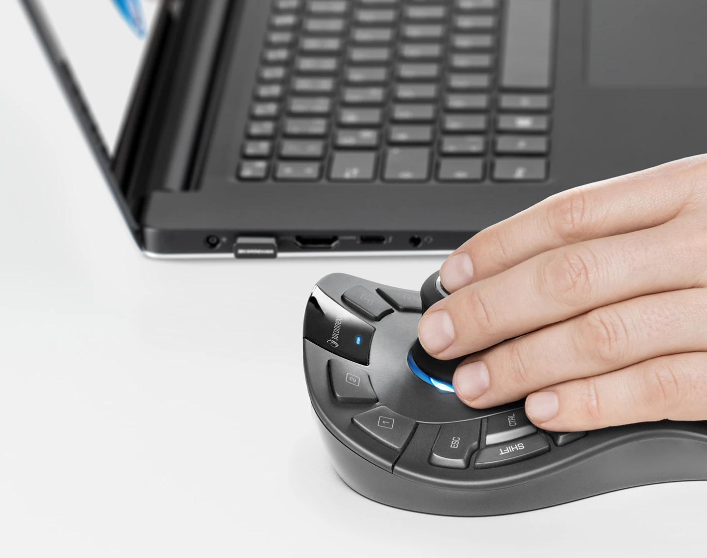 A hand using a 3Dconnexion mouse with a laptop in the background.