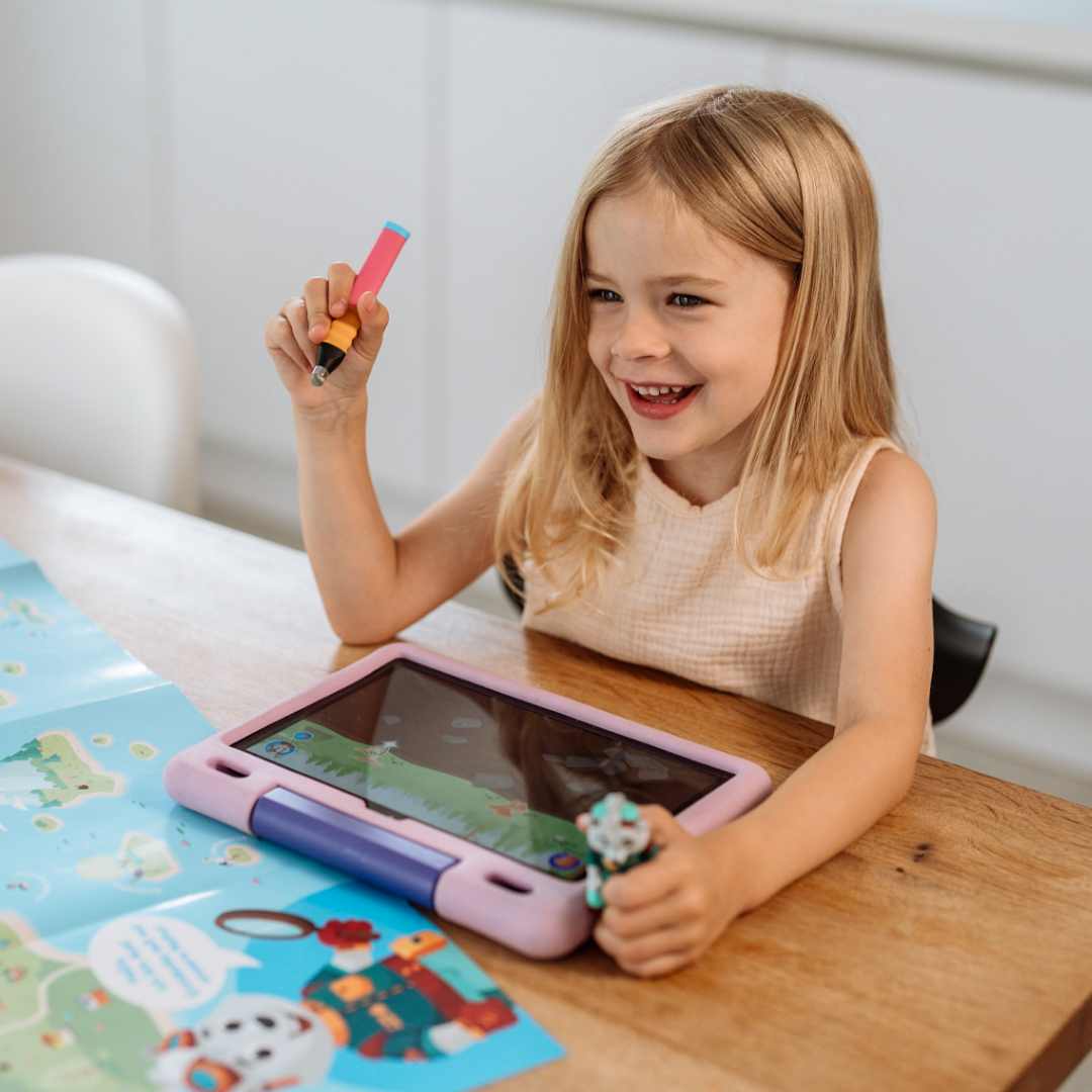 Junges Mädchen mit blonden Haaren lächelt, während es einen Stift und ein Spielzeug hält. Ein Tablet und eine Karte liegen auf dem Tisch.