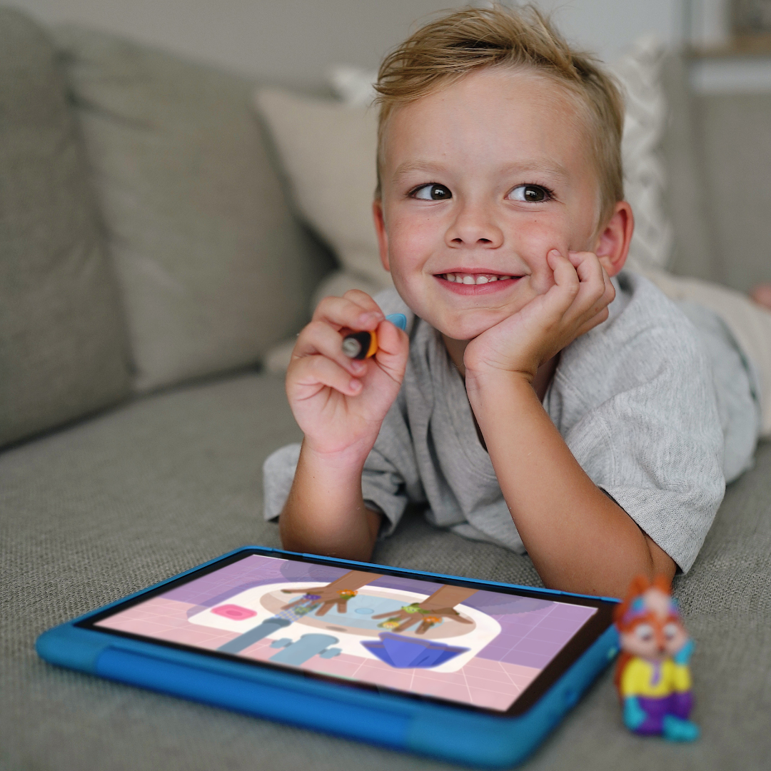 Ein lächelnder Junge liegt auf einer Couch und schaut auf ein Tablet. Er hält einen Stift, ein Spielzeug neben ihm.