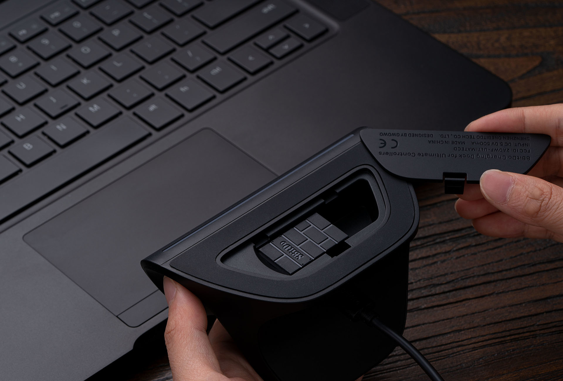 Osoba trzyma czarną stację ładującą obok laptopa. Stacja jest otwierana, ukazując wzór przypominający cegły.