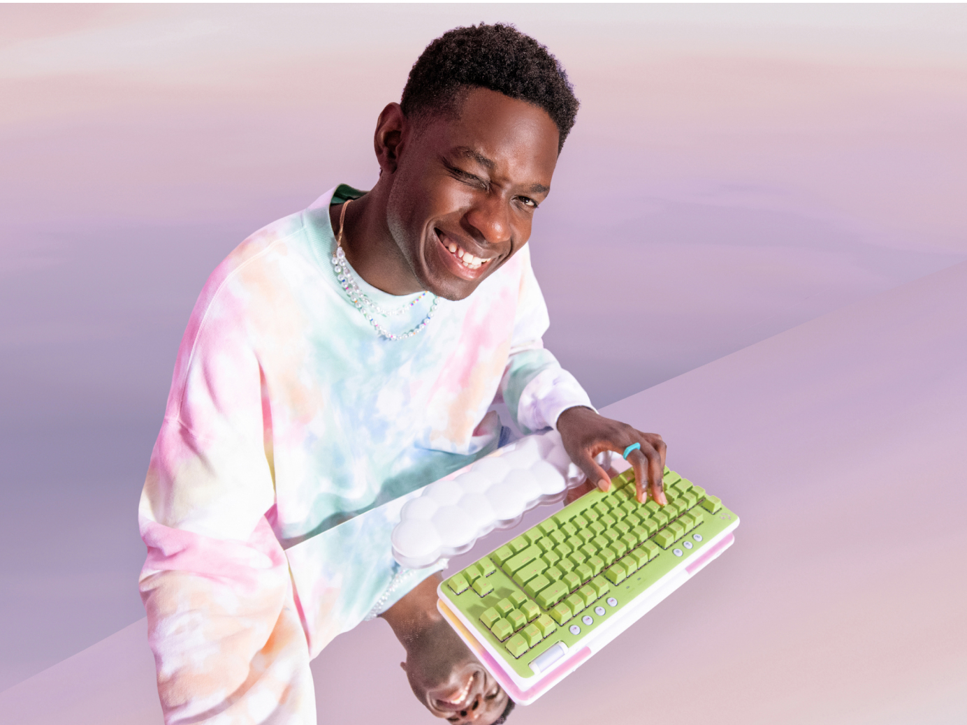 Mężczyzna siedzący przy biurku z klawiaturą i podpórką pod nadgarstek, uśmiechnięty. Fioletowe tło.