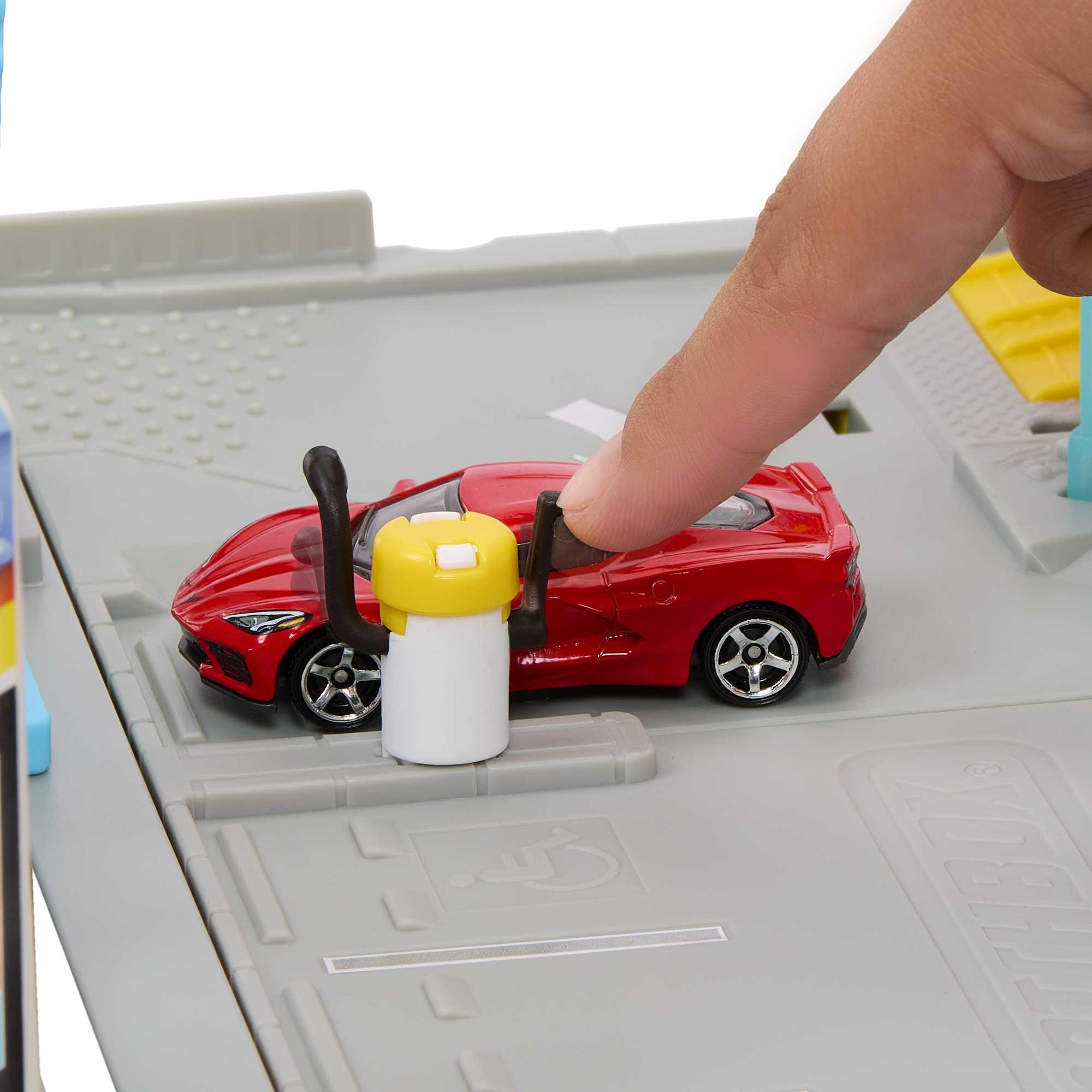 Eine Hand interagiert mit einem roten Spielzeugauto in einer Spielzeuggarage. Eine weiße Zapfsäule ist vorhanden.