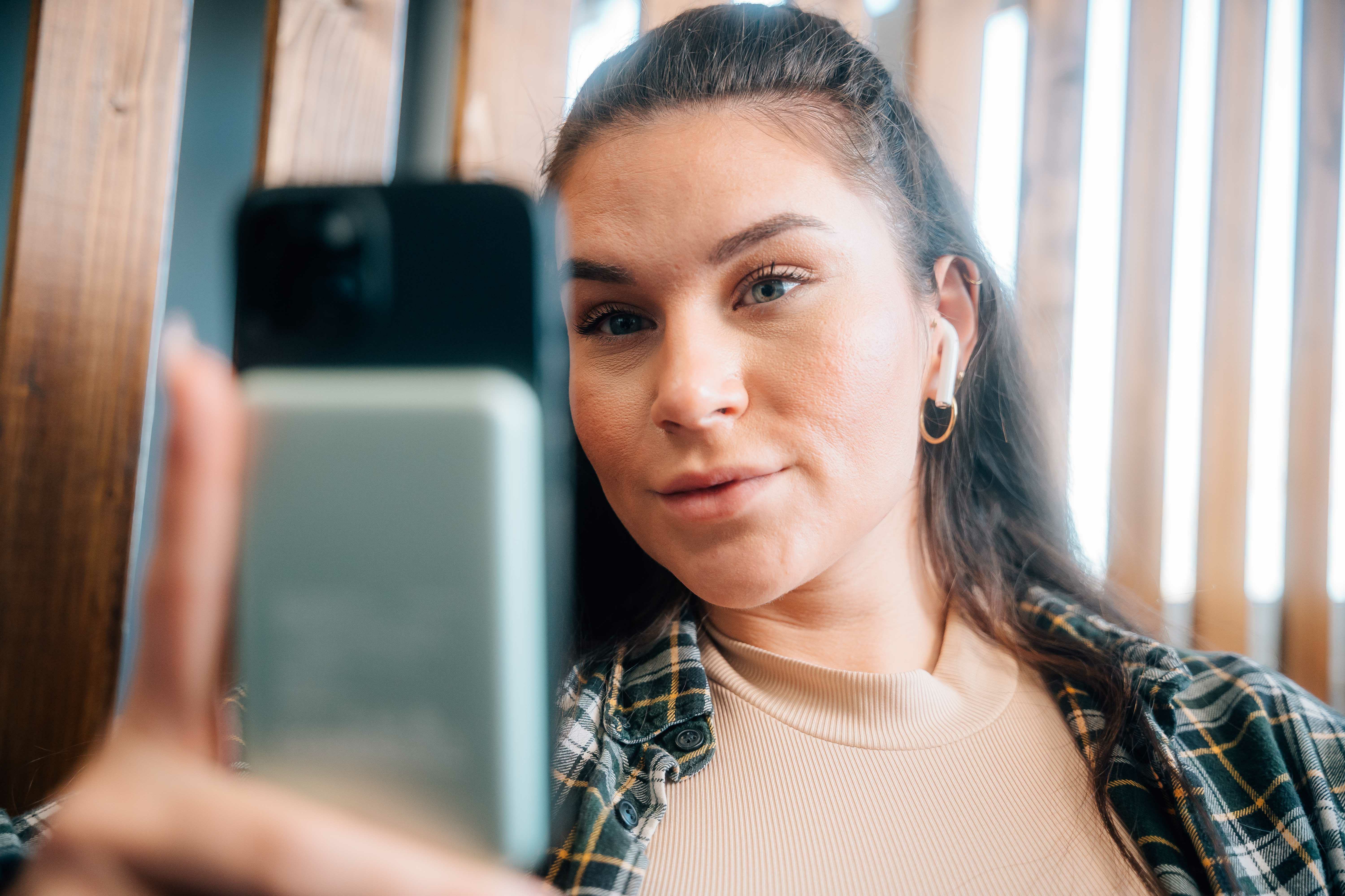 Młoda kobieta robi selfie smartfonem, ubrana w koszulę w kratę i kolczyki.