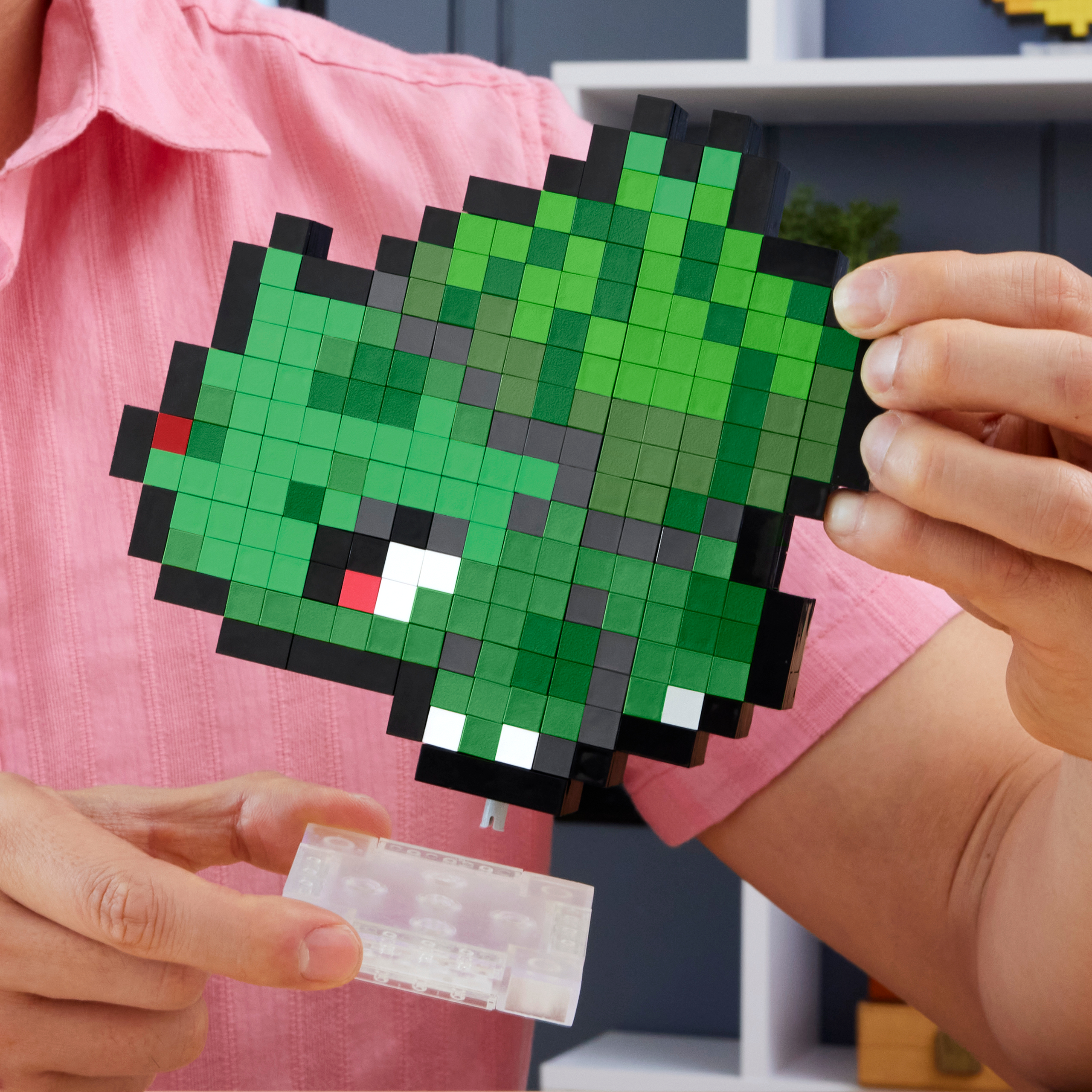 Una persona tiene in mano un drago pixelato verde fatto di blocchi, con una base di plastica trasparente davanti.