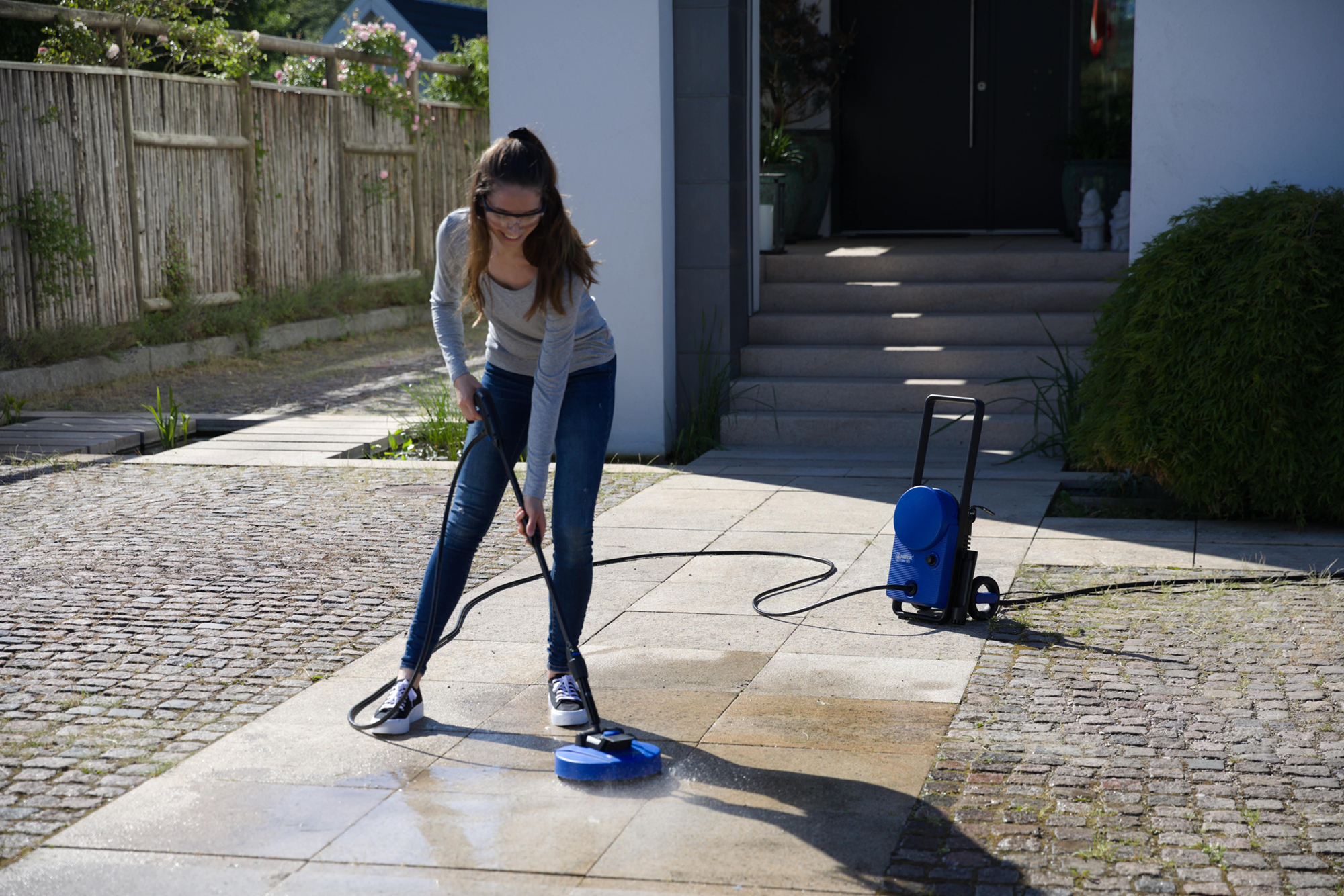 Kobieta czyszcząca brukowany obszar myjką ciśnieniową. Nosi okulary i dżinsy.