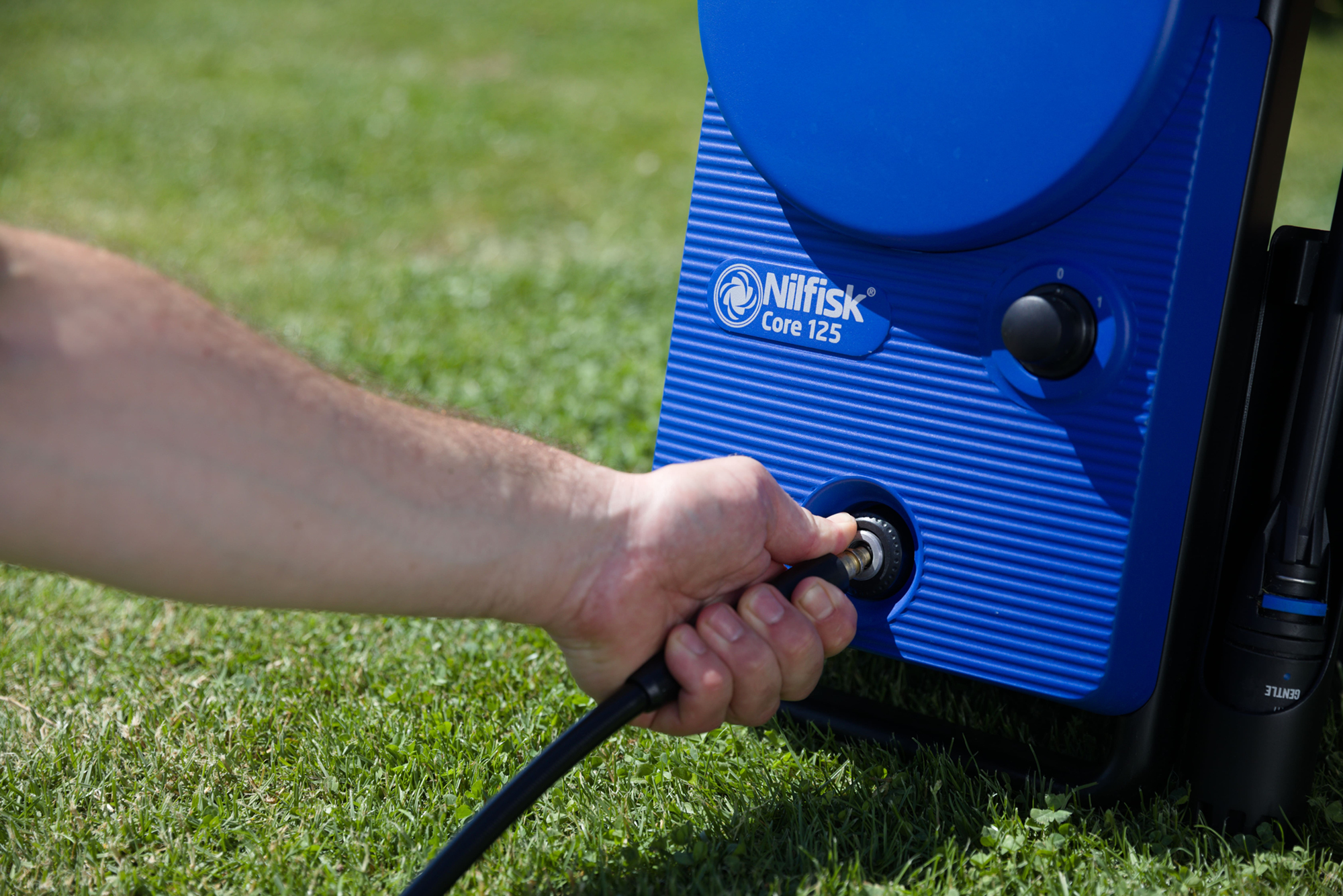 Ręka osoby podłączająca czarny wąż do niebieskiej myjki ciśnieniowej, na zielonej trawie.