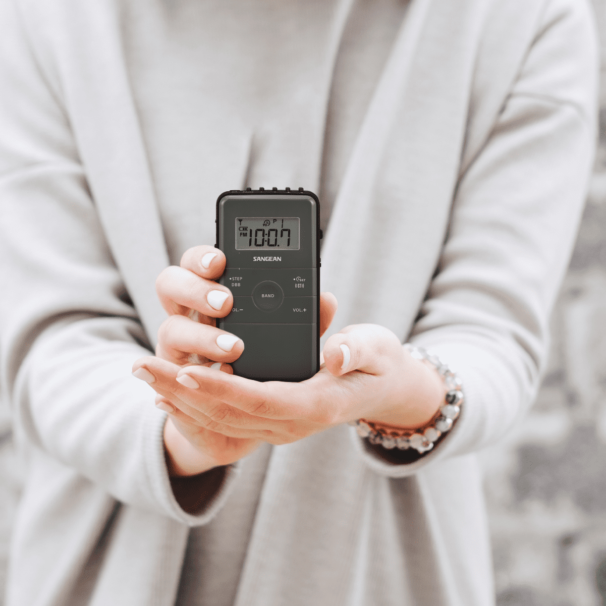 Osoba trzymająca ciemnoszare radio Sangean, pokazujące godzinę, na neutralnym tle.