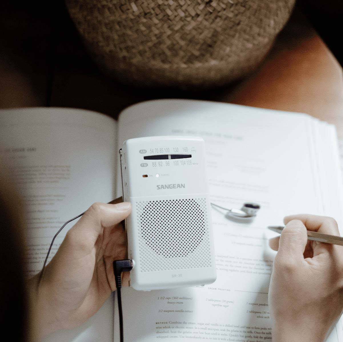 Białe radio z kratką głośnika i pokrętłem. Ręce trzymają radio obok książki.