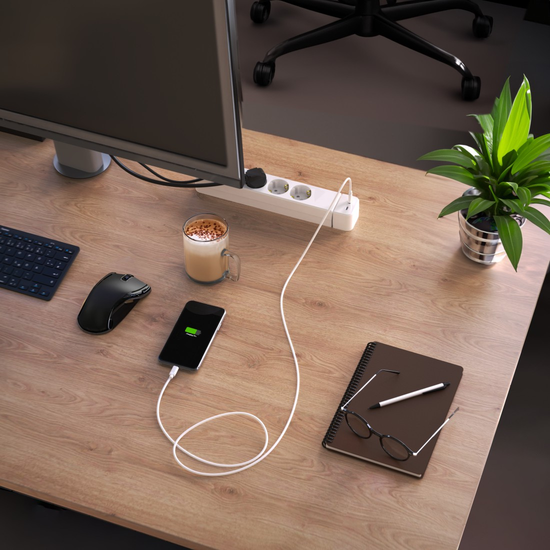 Biurko z monitorem, myszką, klawiaturą, telefonem w ładowaniu, kawą, notatnikiem i rośliną.