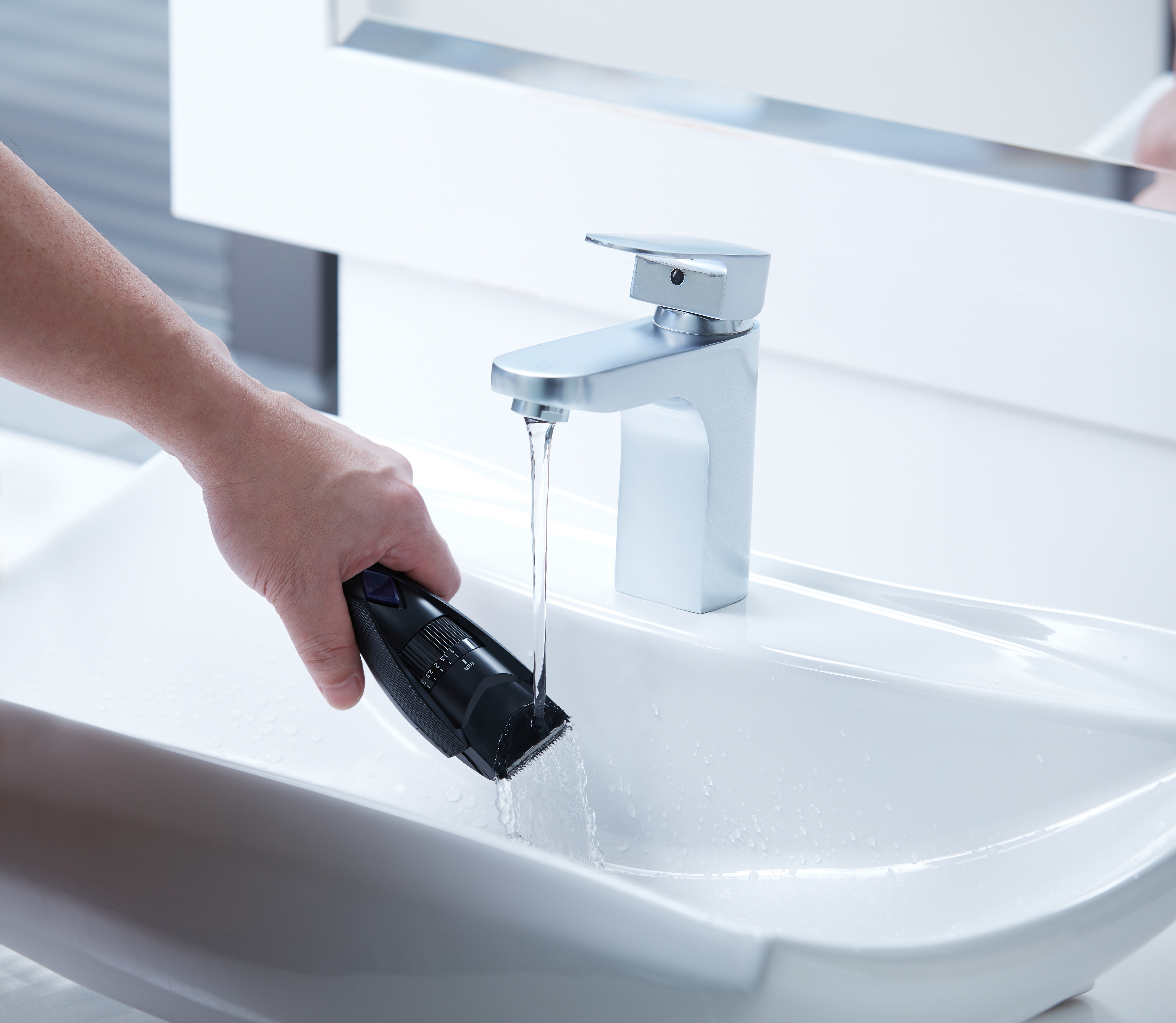 Una mano sostiene una recortadora negra sobre un lavabo blanco con agua corriente y un espejo.