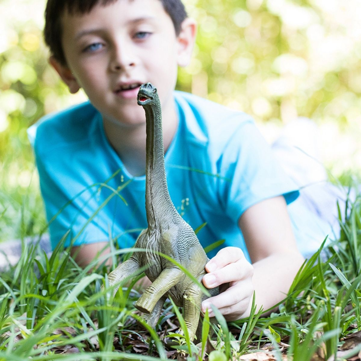 Ein Junge hält einen Spielzeug-Dinosaurier im Gras. Der Dinosaurier ist grün.