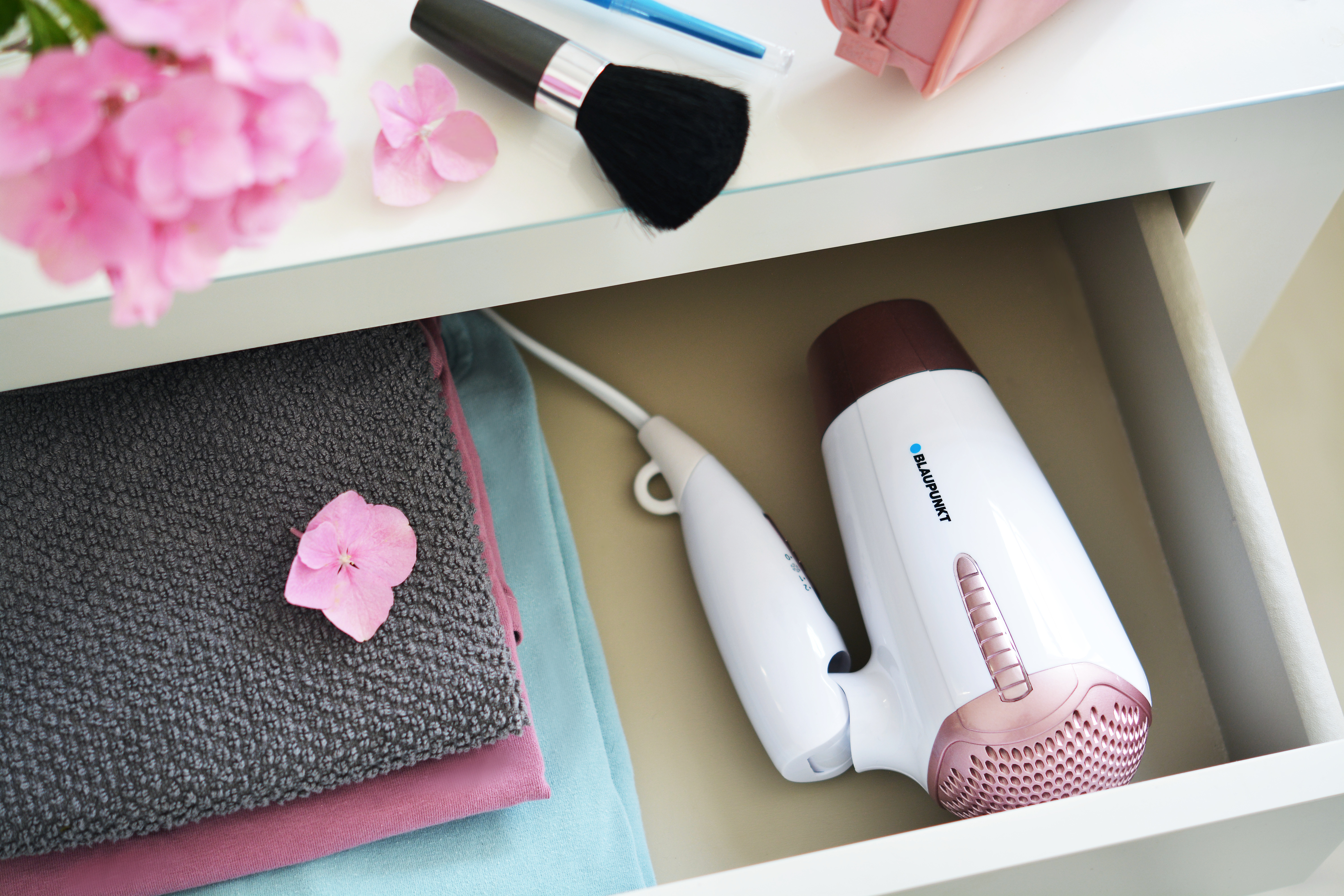 Otwarta szuflada z suszarką, ręcznikiem, szczotką i kwiatami.