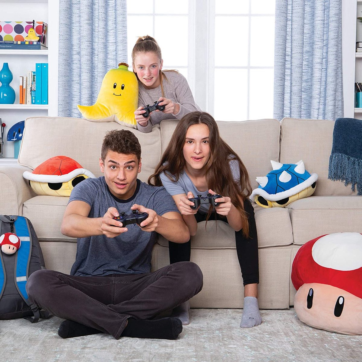 Tre giovani che giocano ai videogiochi su un divano beige. Controller di gioco, cuscini e uno zaino.