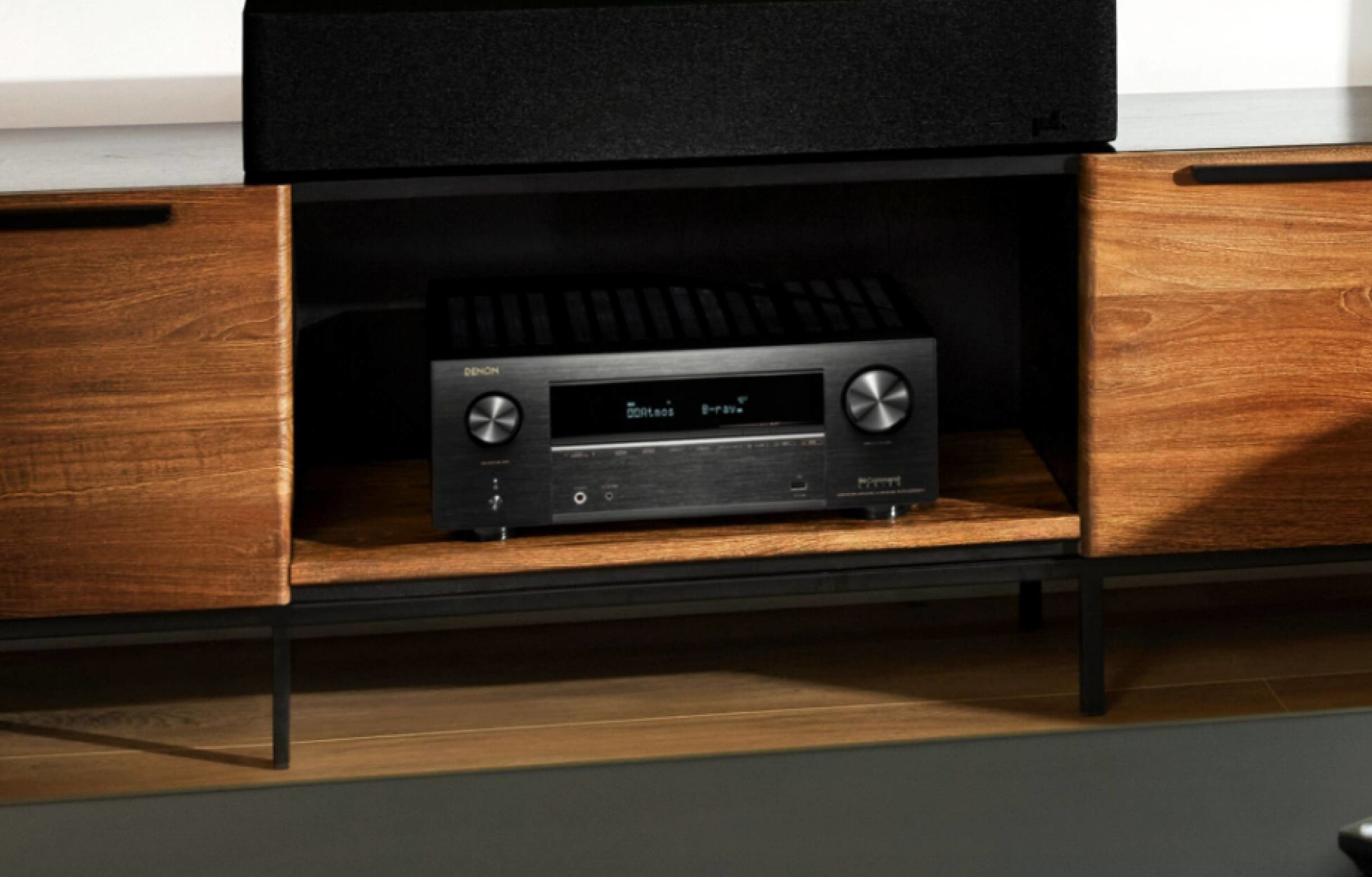Receptor de audio negro en un estante, con paneles de madera y una barra de sonido arriba.