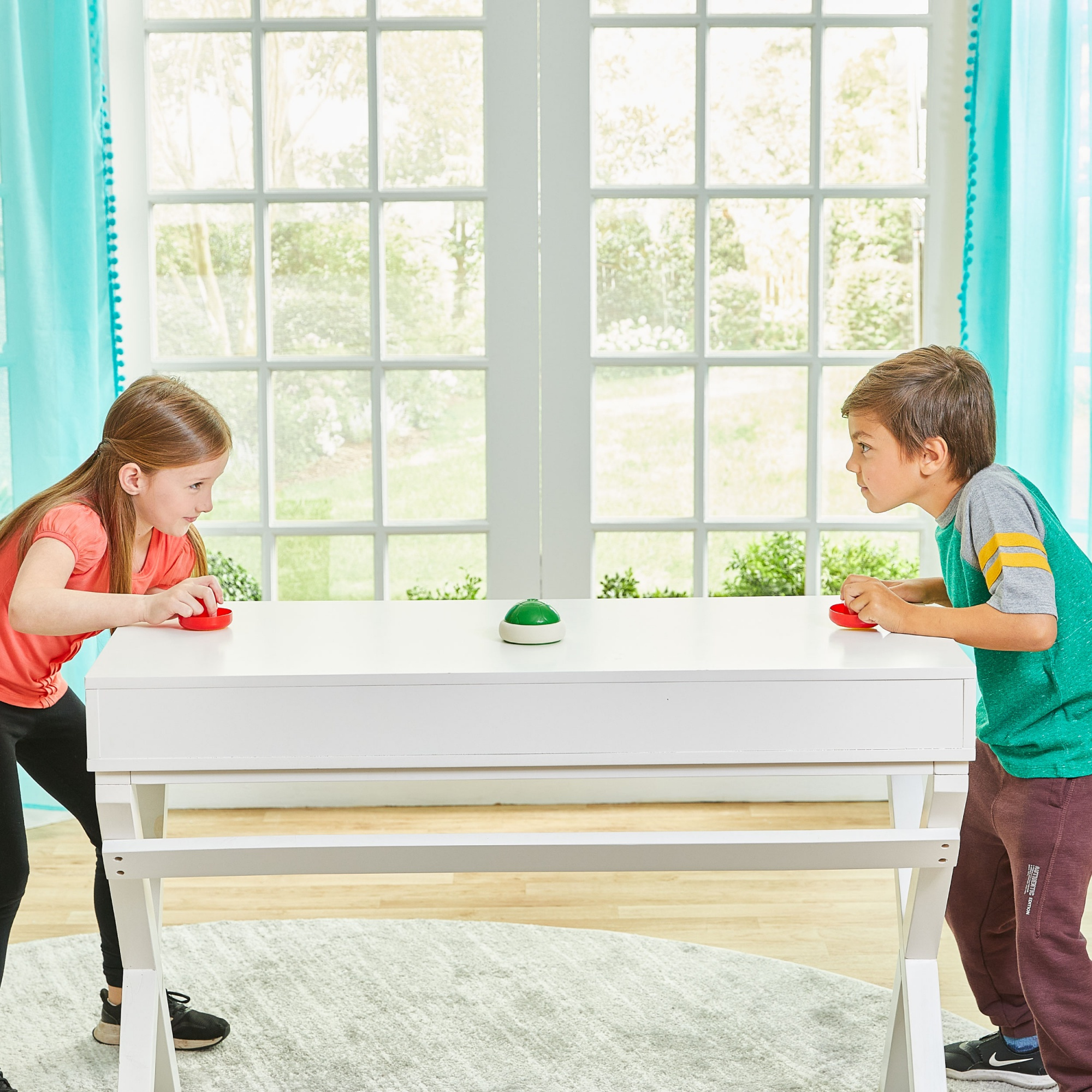 Dwoje dzieci grających w grę stołową. Biały stół, czerwone i zielone przyciski. Okna w tle.