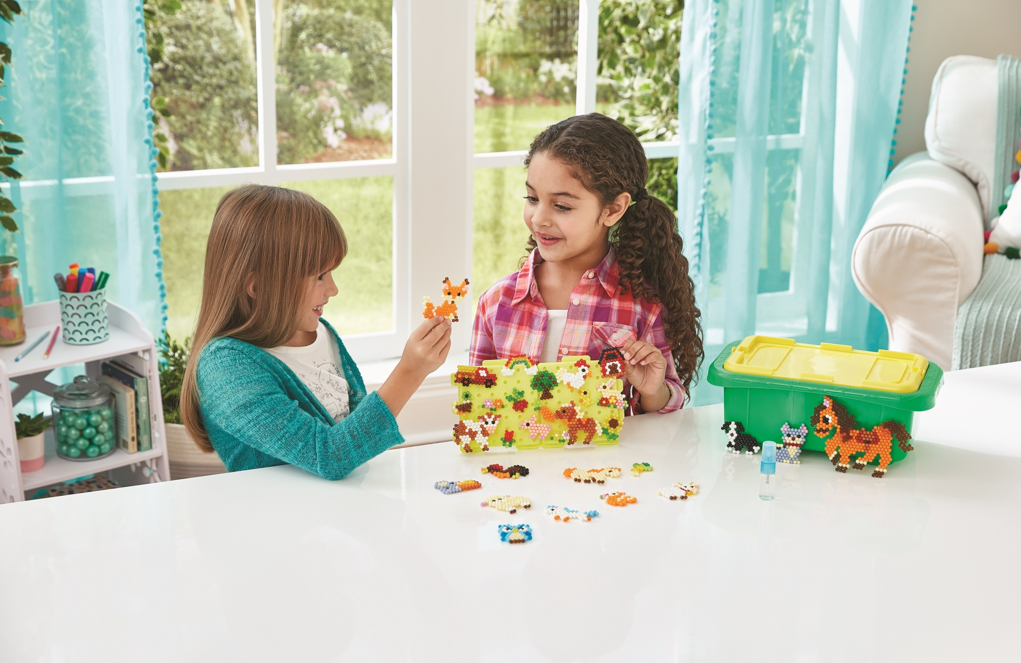 Dos niñas jugando con cuentas de colores y una caja de juguetes, en una mesa blanca cerca de una ventana.