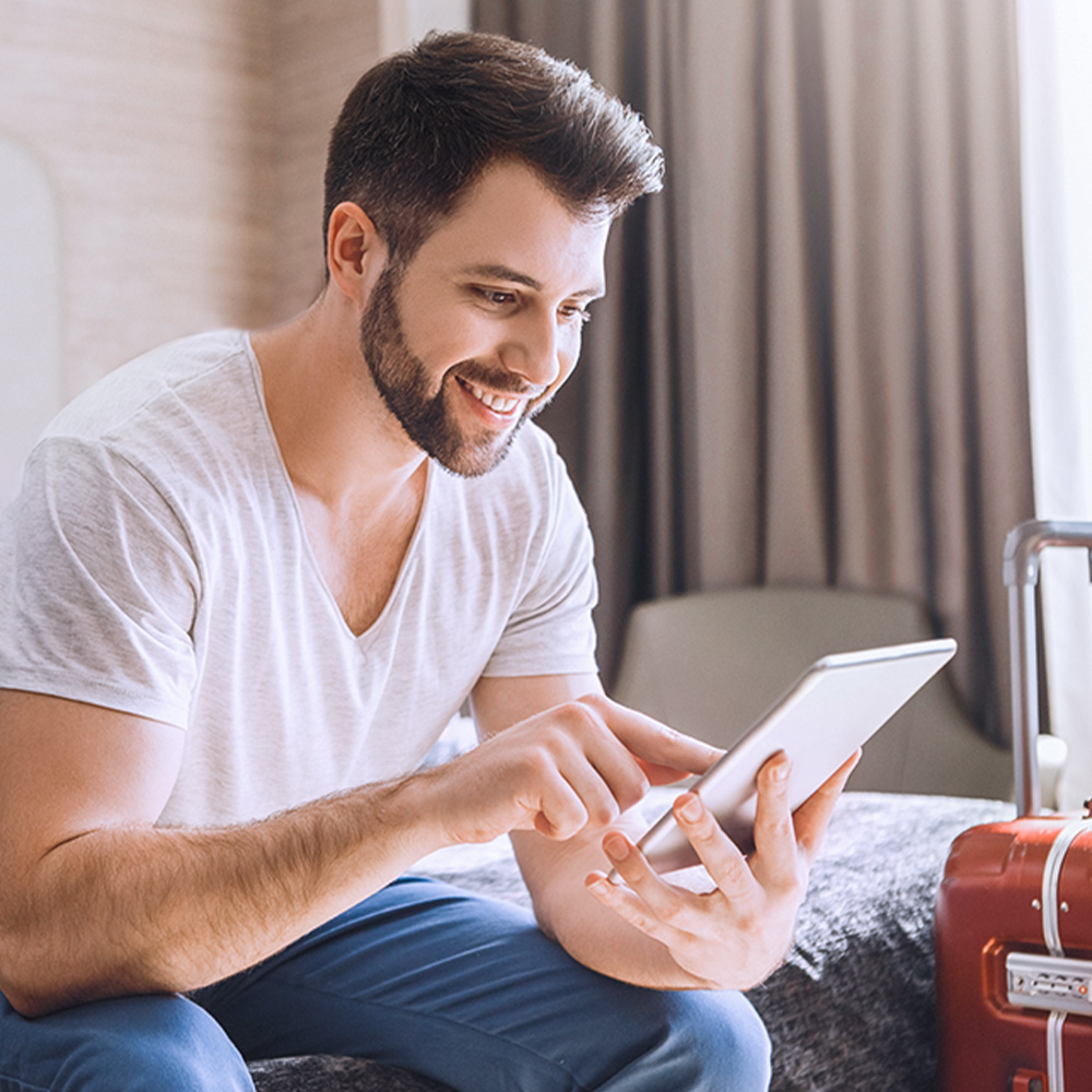 Uśmiechnięty mężczyzna korzystający z tabletu. Na łóżku czerwona walizka.