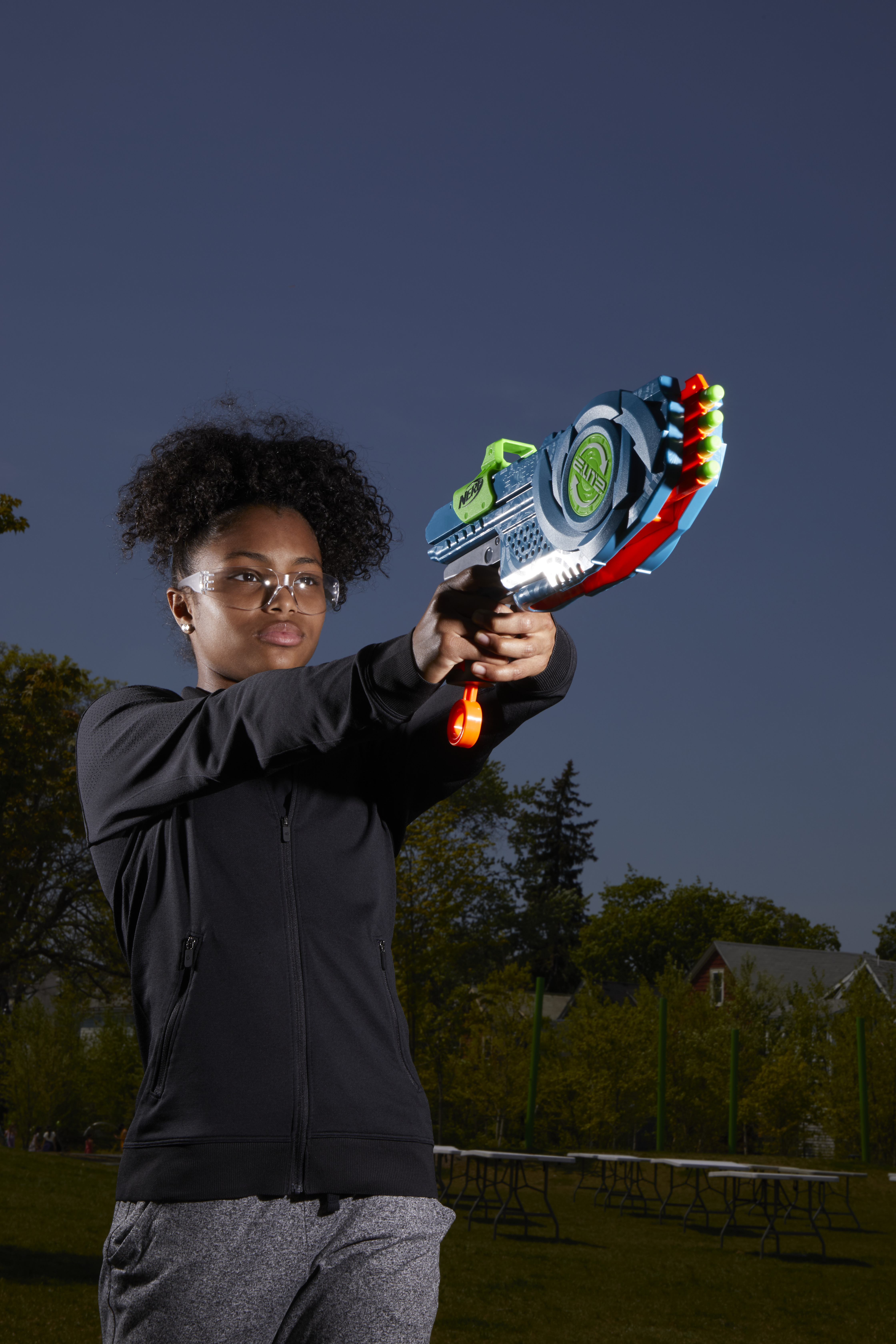 Una ragazza con gli occhiali che tiene una pistola giocattolo, puntando verso un cielo blu scuro al tramonto.