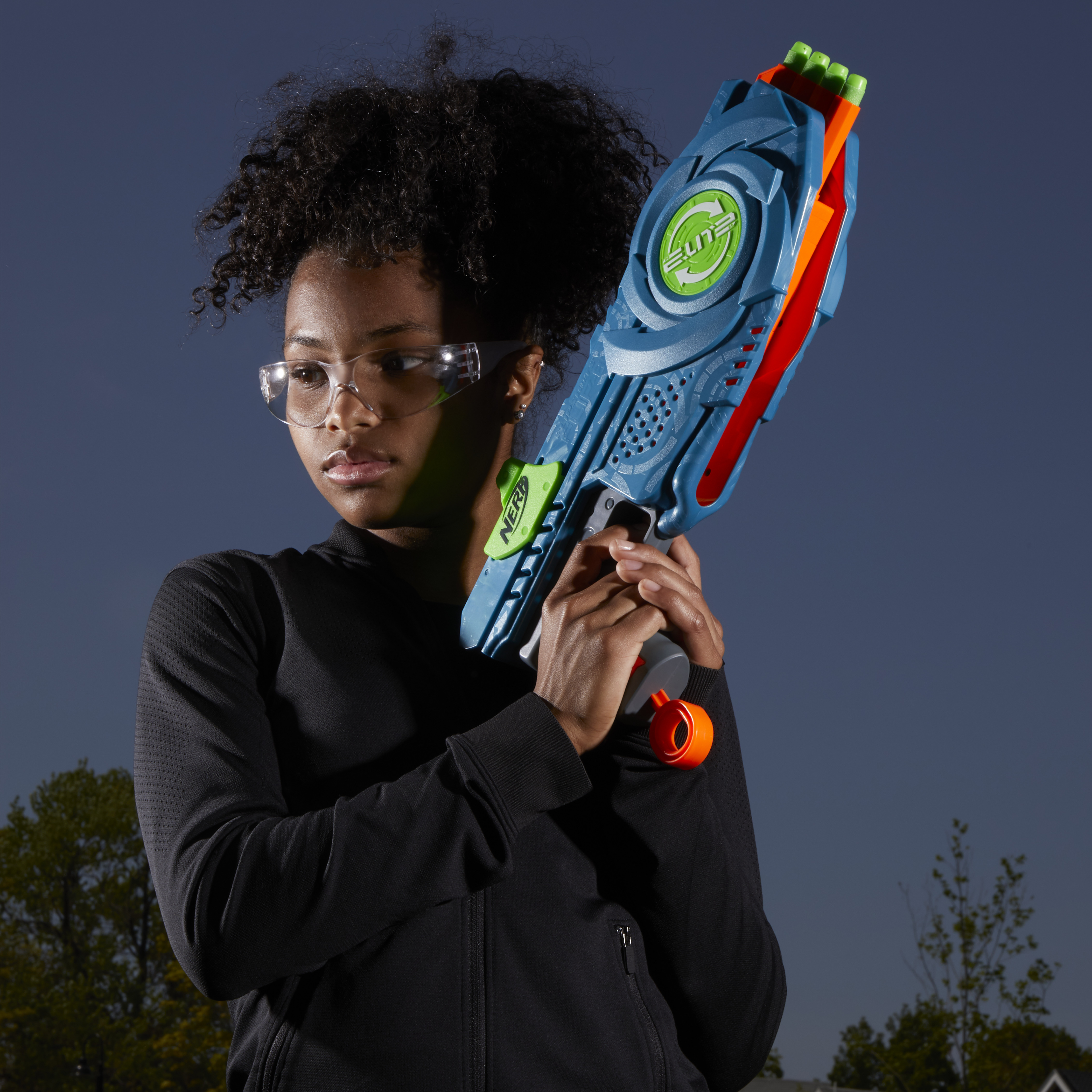 Una ragazza con occhiali protettivi tiene in mano una pistola Nerf blu davanti a un cielo blu scuro.