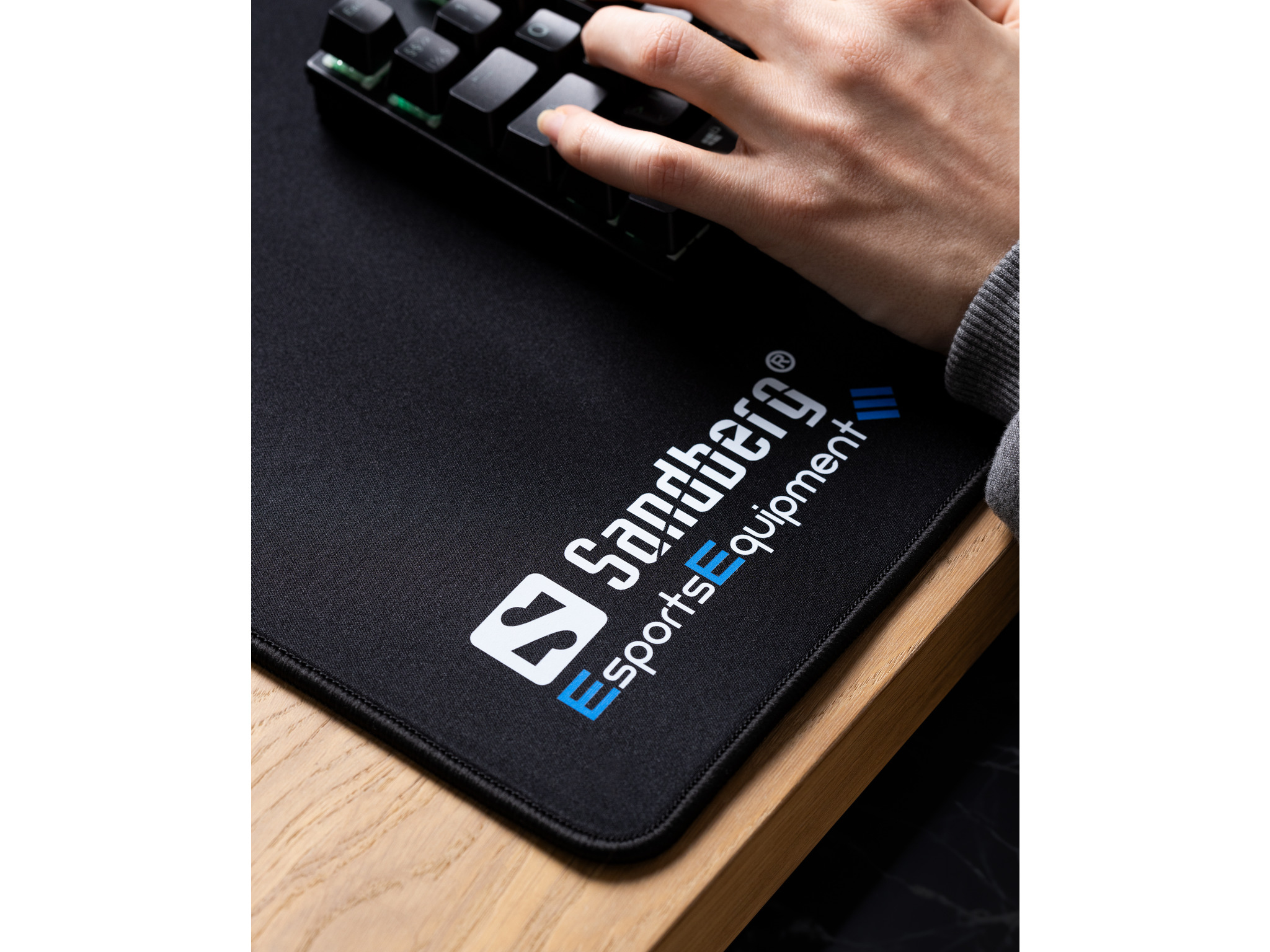 Un tapis de souris noir avec le logo Sandberg et une main utilisant un clavier noir.