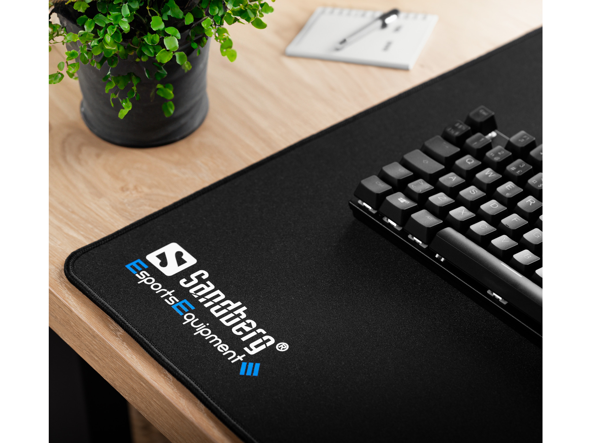 Tapis de souris noir avec clavier et plante sur un bureau en bois. Logo Sandberg sur le tapis de souris.