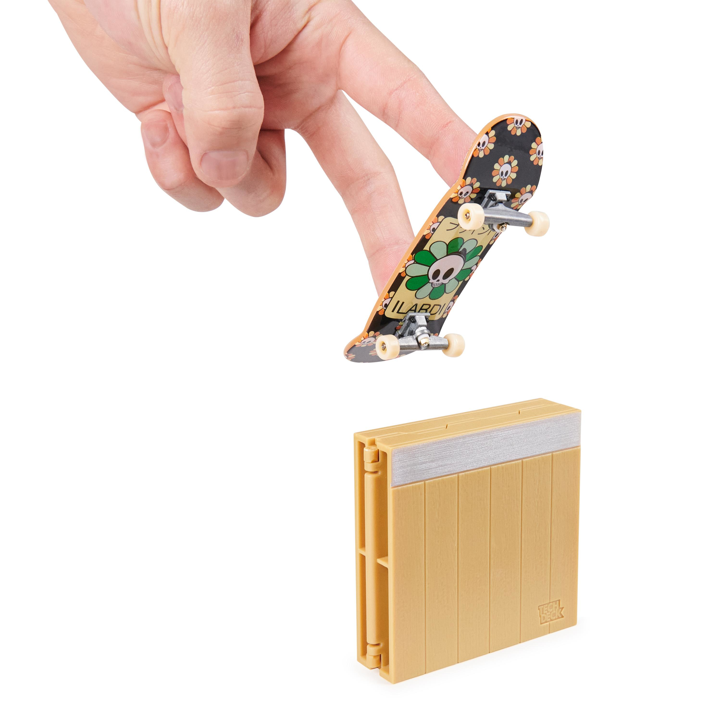Finger halten ein Spielzeug-Skateboard über einer gelben Rampe. Das Skateboard hat ein Blumendesign.