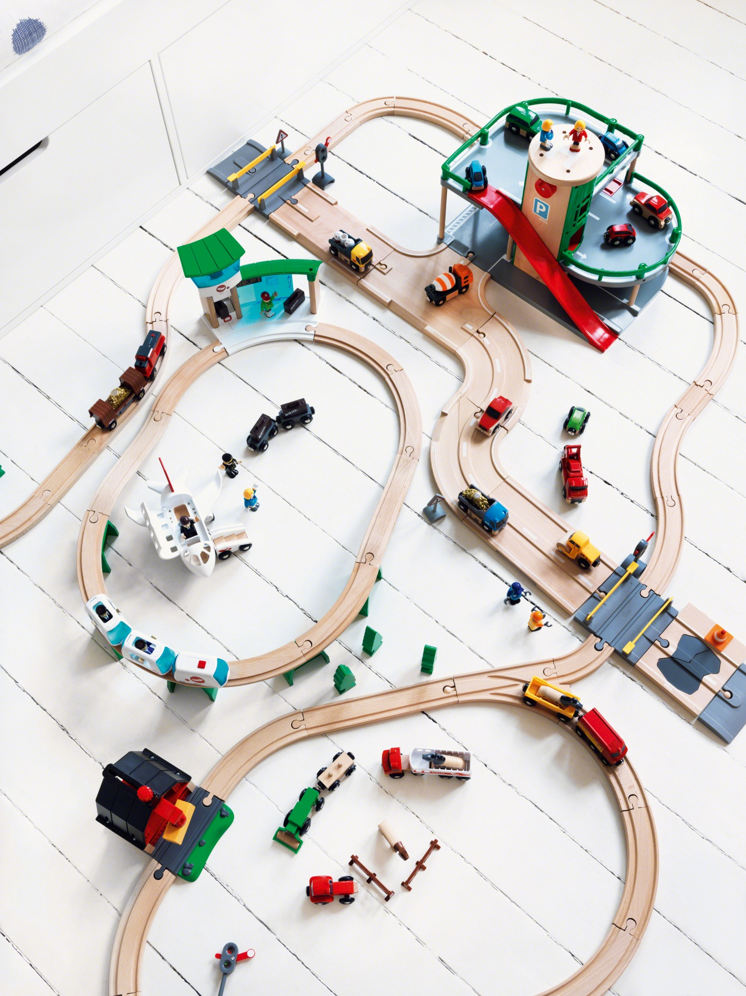 Set di trenini giocattolo con binari, edifici, veicoli e figure su una superficie bianca, visto dall'alto.