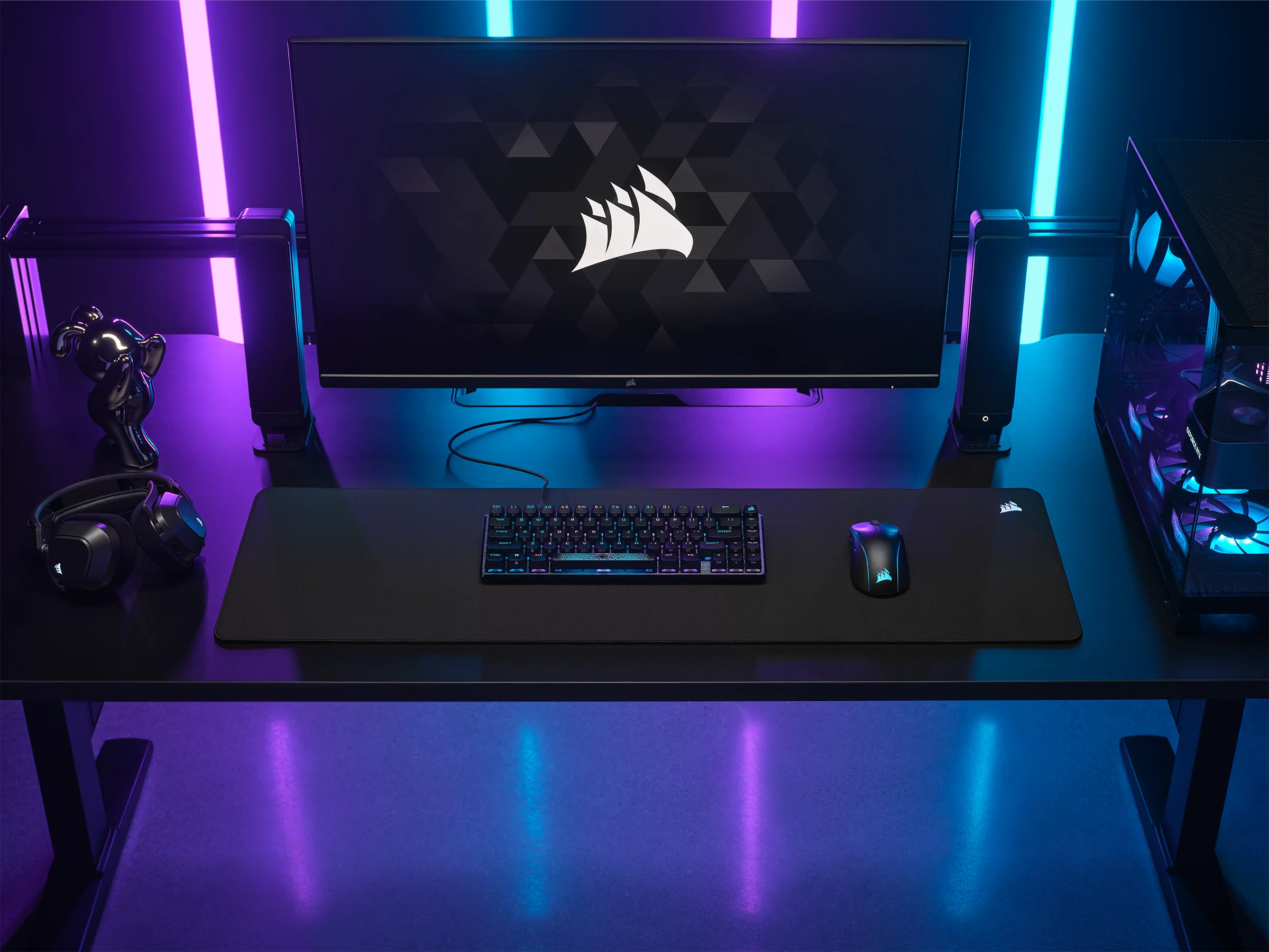 Konfiguracja biurka z monitorem, klawiaturą, myszą, głośnikami i obudową komputera. Niebieskie/fioletowe oświetlenie.