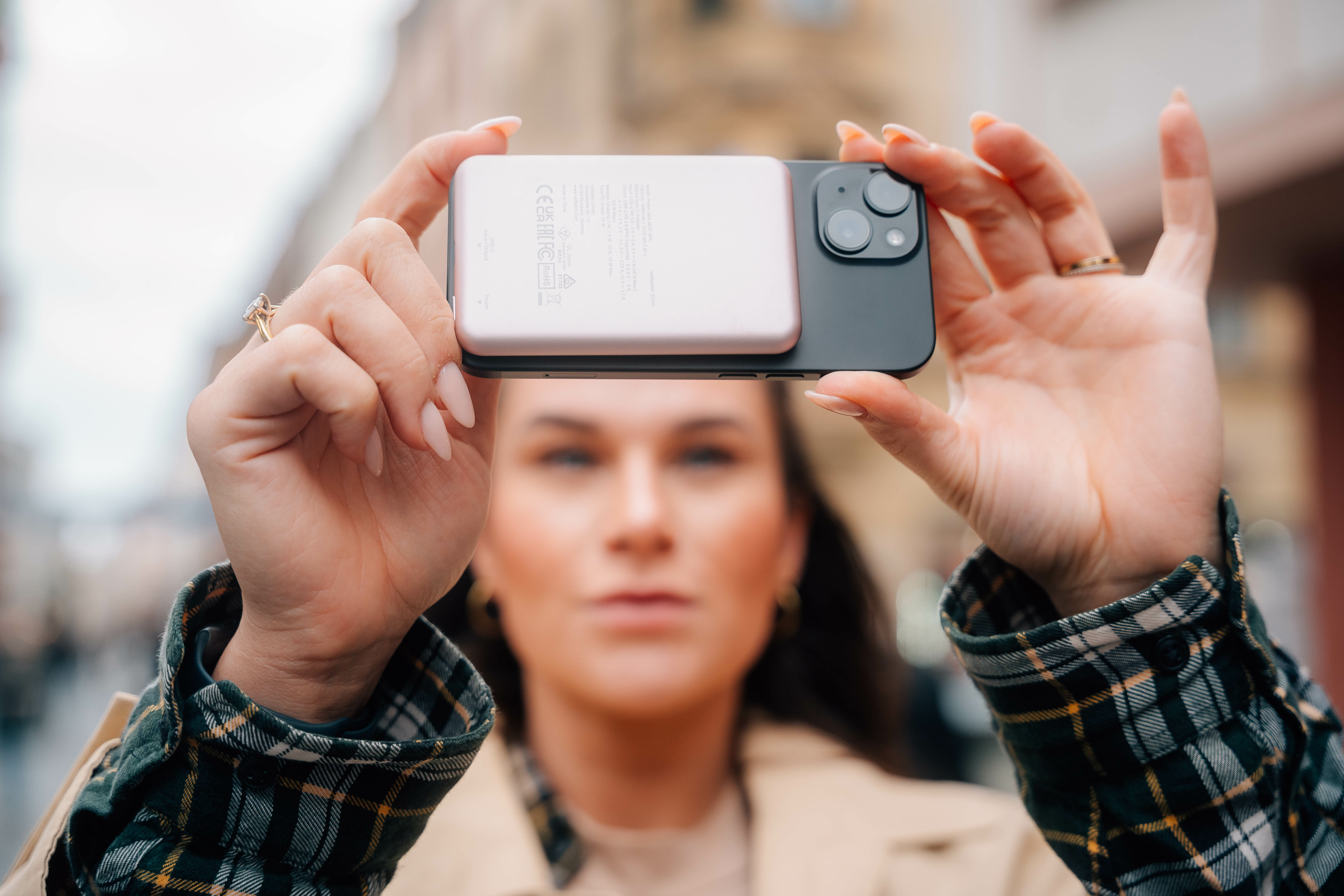 Kobieta trzymająca telefon i power bank, rozmyte tło budynków.