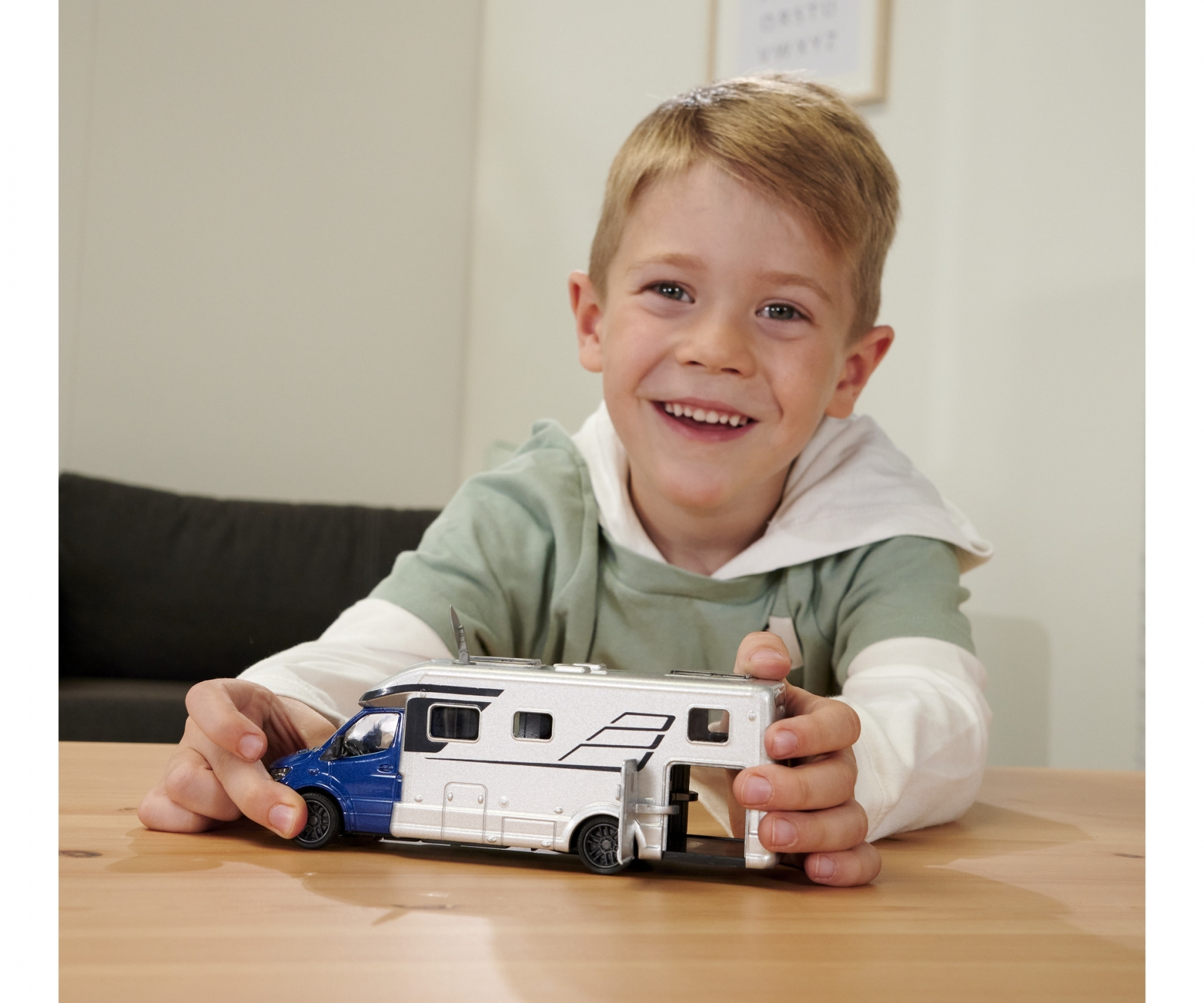 Uśmiechnięty chłopiec trzyma srebrną i niebieską zabawkę kampera. Białe tło, wesoła scena.