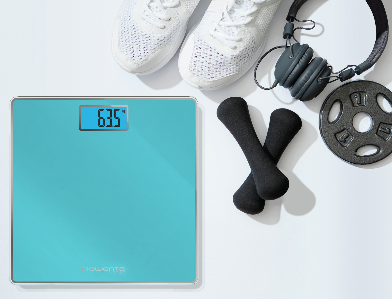 Waga cyfrowa pokazująca 63,5 kg, obok białych butów sportowych, czarnych hantli i słuchawek.