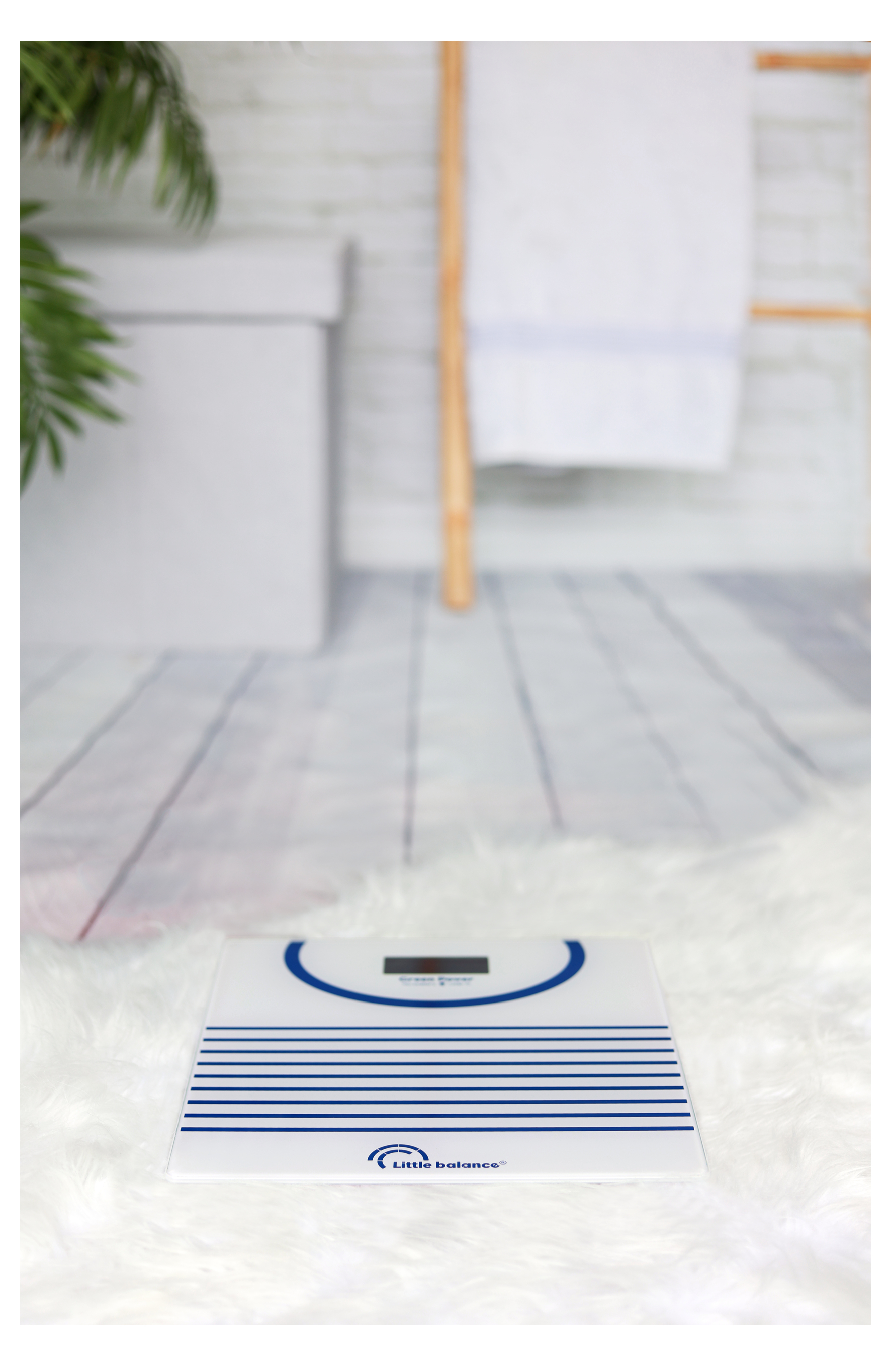 Biała waga łazienkowa na puszystym, białym dywanie. W tle wieszak na ręczniki i roślina.