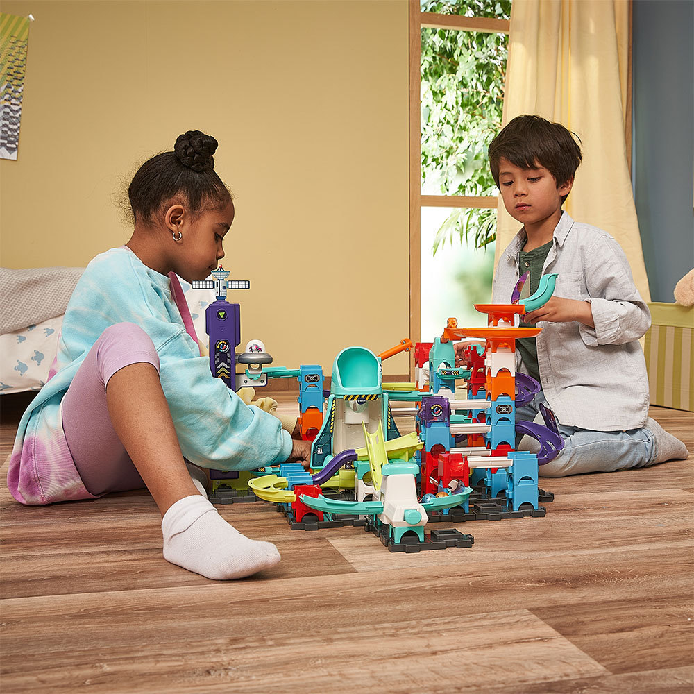 Zwei Kinder spielen mit einer Spielzeug-Autobahn, mit einer hellblau-orangenen Strecke.