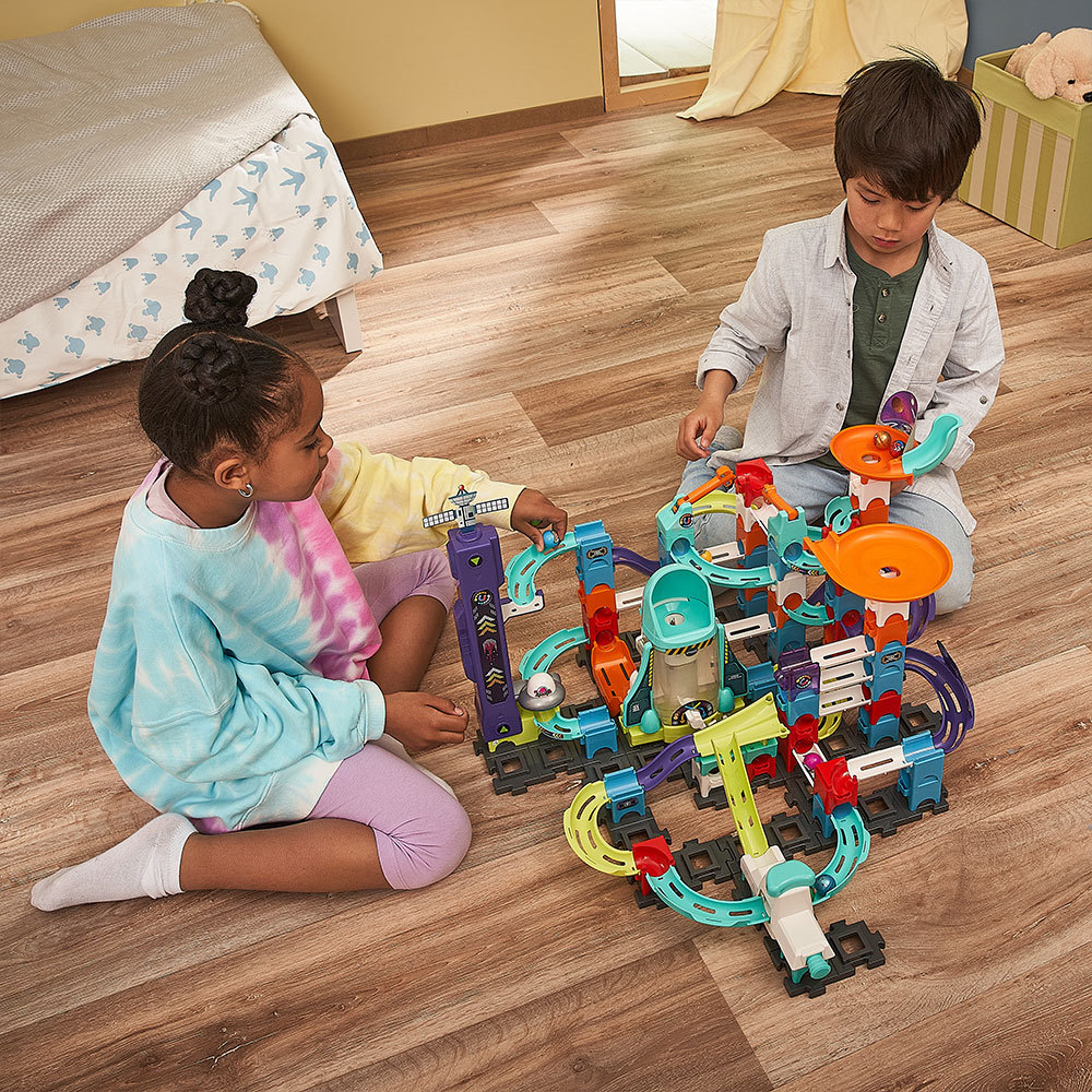 Zwei Kinder spielen mit einem Spielzeug-Konstruktionsset auf einem Holzboden. Verschiedene farbige Teile.
