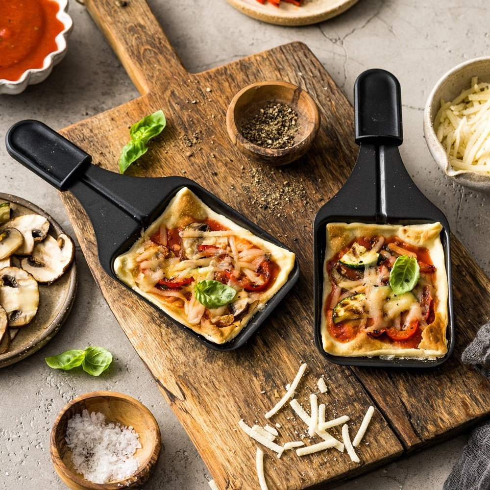 Dwie mini pizze w czarnych patelniach na drewnianej desce. Składniki takie jak pieczarki, ser i bazylia.