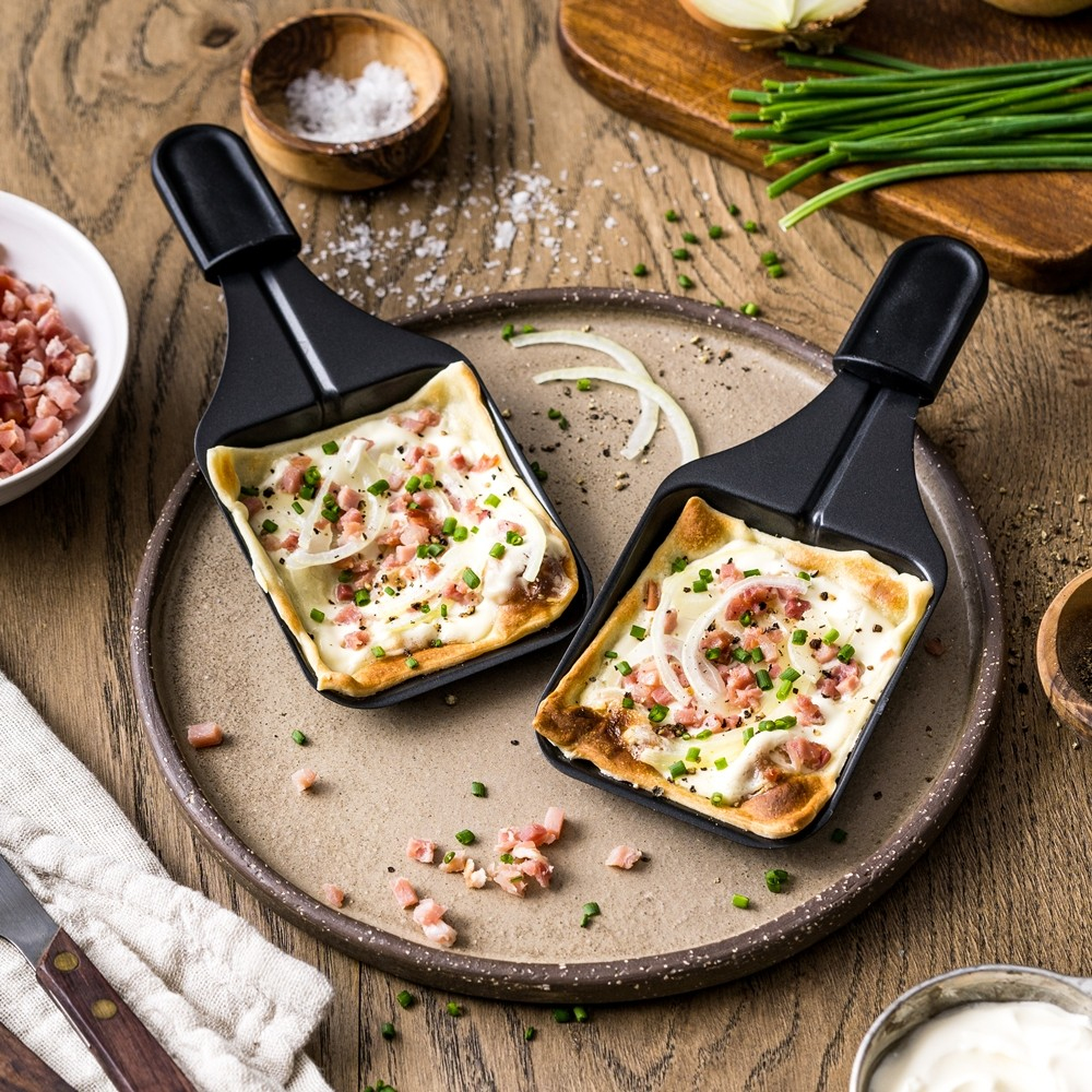 Dwie patelnie do raclette z roztopionym serem, cebulą i szynką na drewnianym stole ze składnikami.