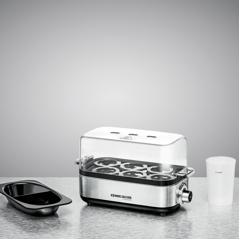 Garnek do gotowania jajek z przezroczystą pokrywą, czarną tacą i białą miarką. Tło jest szare.