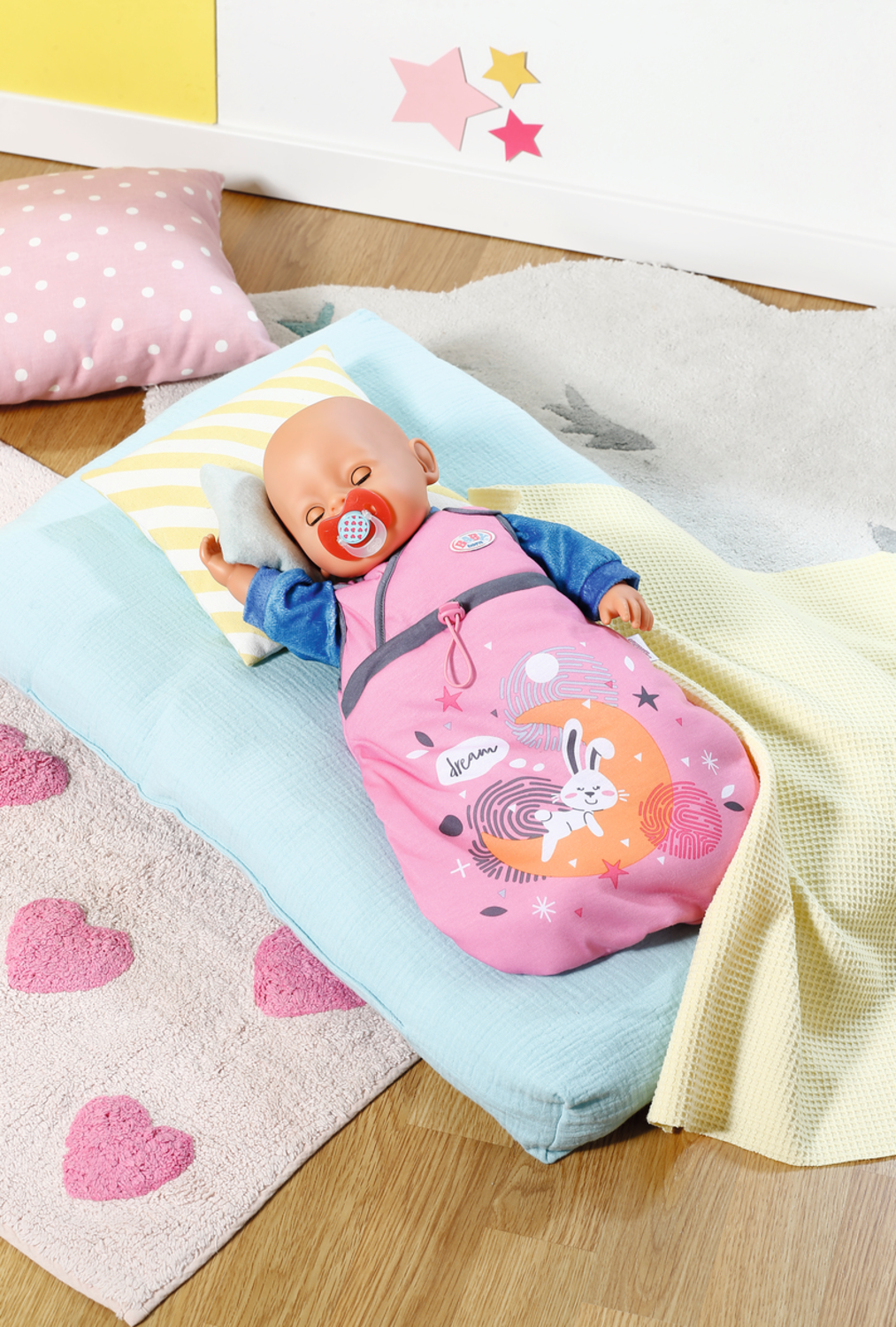 Muñeca bebé durmiendo en un saco de dormir rosa sobre una manta azul. También se ven almohada y alfombra.