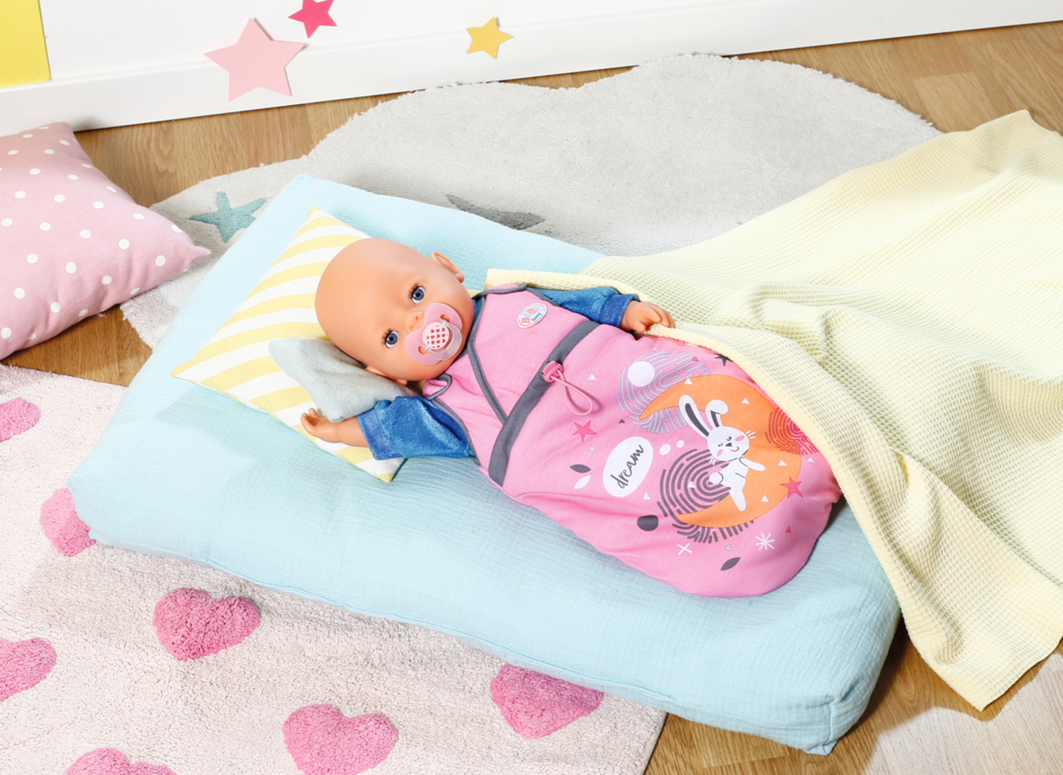 Muñeca bebé en saco de dormir rosa sobre almohada azul, con manta y juguetes.