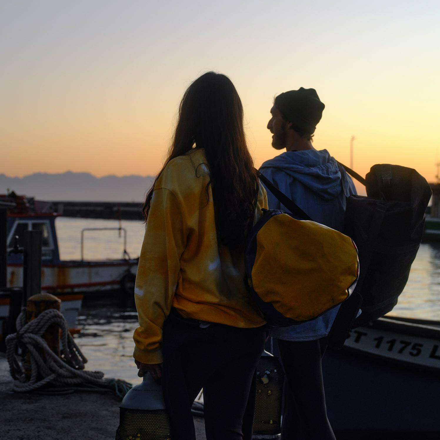 Dwie osoby stojące w pobliżu łodzi o zachodzie słońca. Jedna w żółtym, druga w niebieskim, patrząca na wodę.