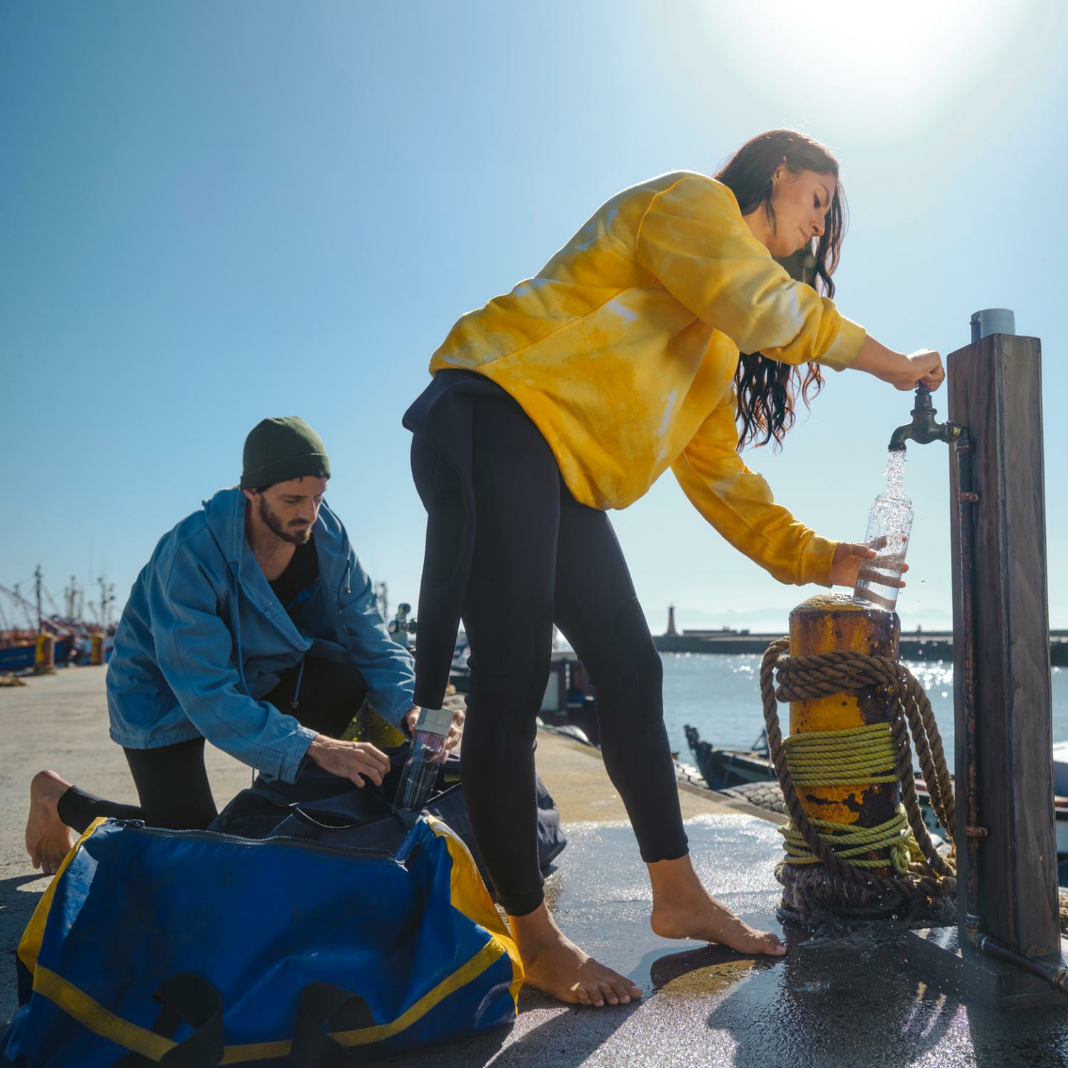 Dwie osoby napełniają butelki z wodą z kranu na pomoście nad morzem w słońcu.