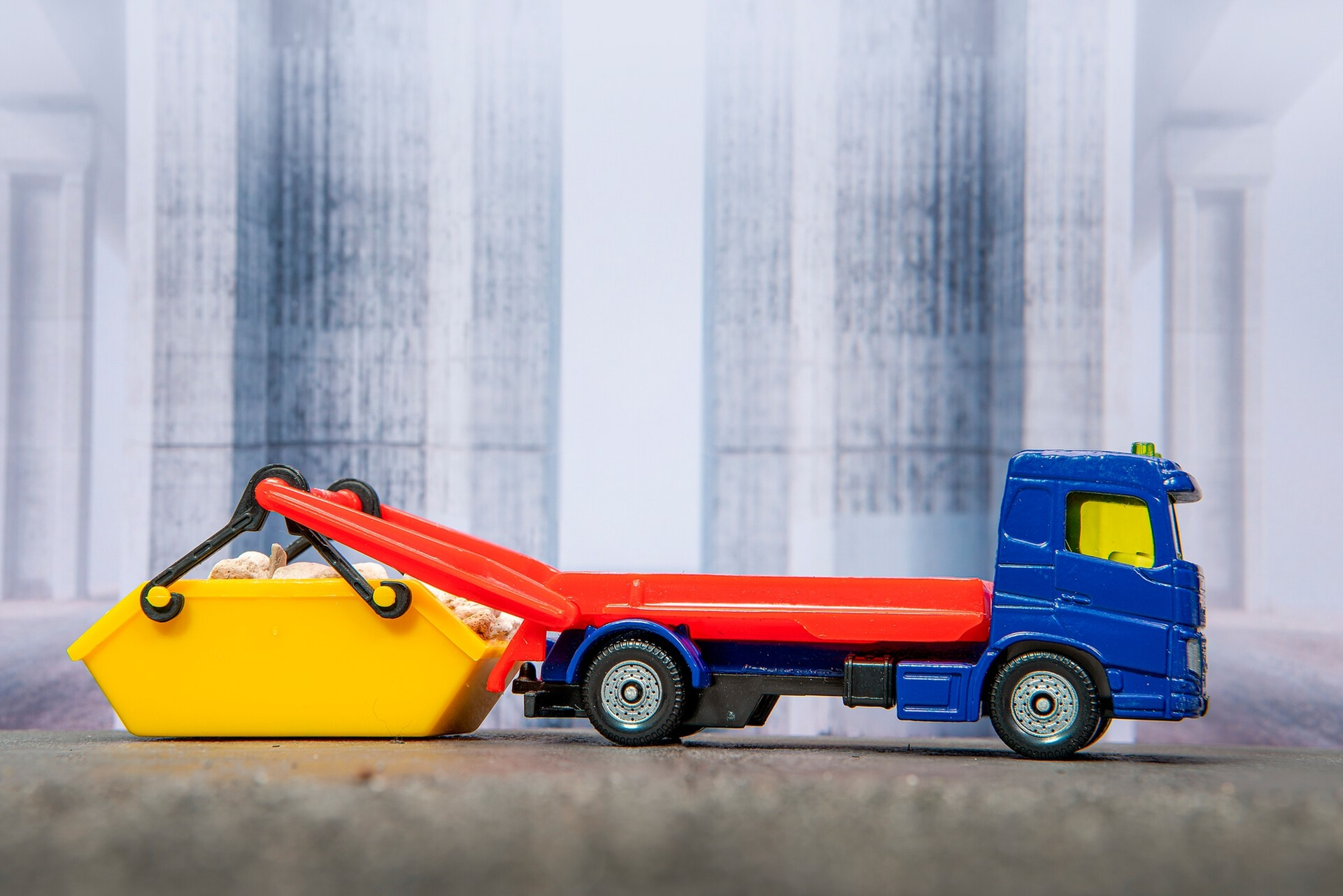 Ein blau-roter Spielzeug-LKW mit einem gelben Container, der angehoben wird. Der Hintergrund ist verschwommen.