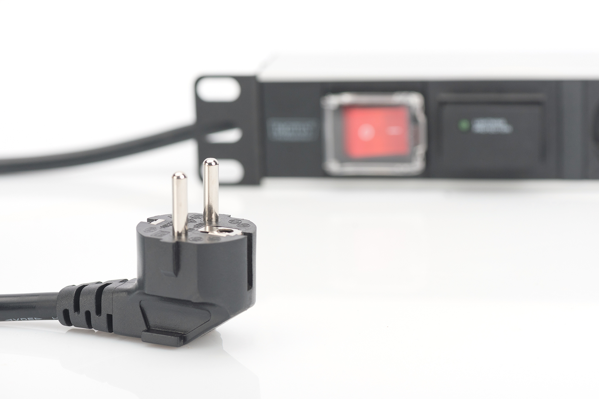 Czarna wtyczka z dwoma metalowymi bolcami na pierwszym planie. Listwa zasilająca z czerwonym przełącznikiem w tle.