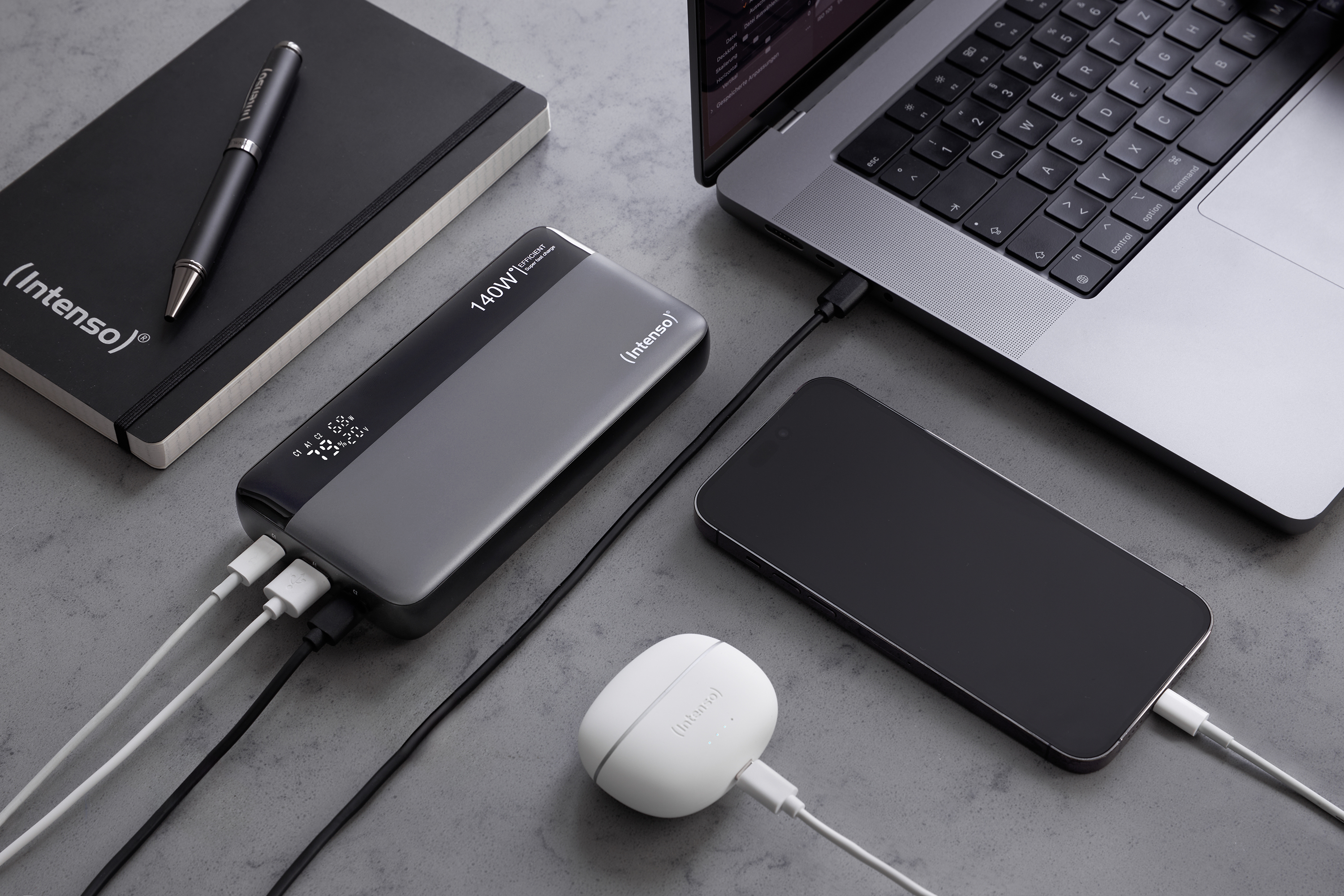 Banco de energía con dispositivos conectados: portátil, teléfono y auriculares. También hay un bolígrafo y un cuaderno.