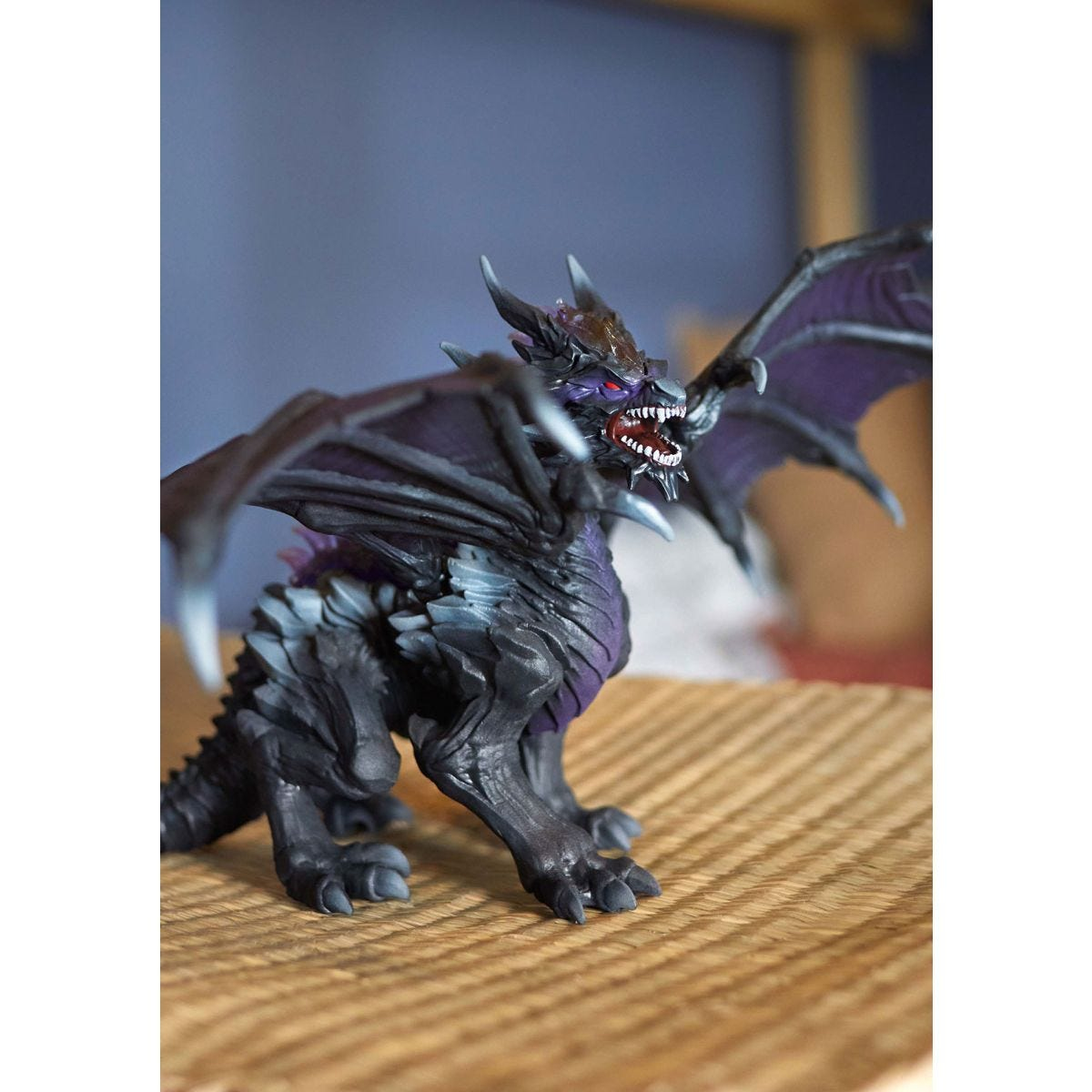 Un giocattolo a forma di drago nero e viola. È in piedi su un tappetino intrecciato, con le ali spiegate. Lo sfondo è sfocato.